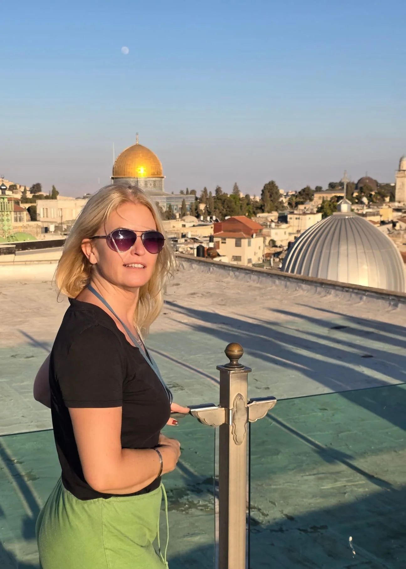 A woman with blonde hair wearing sunglasses, a black t-shirt, and green pants, standing on a rooftop with the skyline of Jerusalem in the background, including the Dome of the Rock with its golden dome.