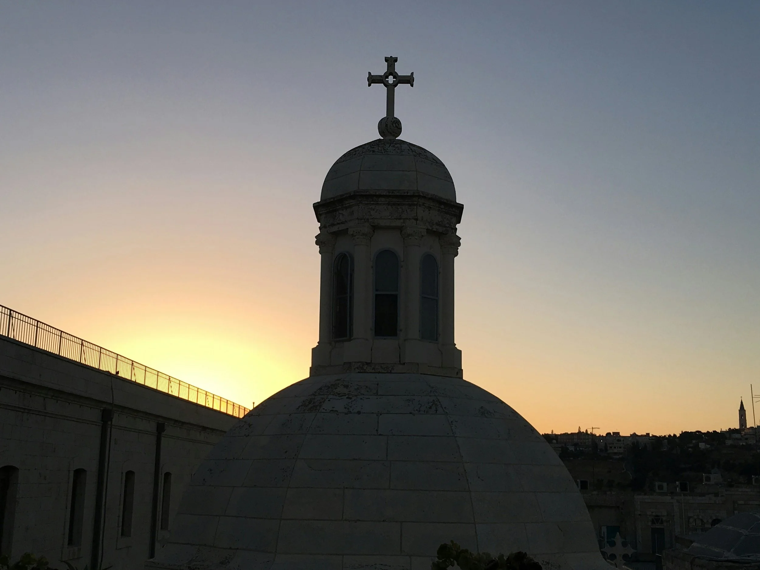 Church tower with a cross silhouetted against a glowing Jerusalem sunset sky.