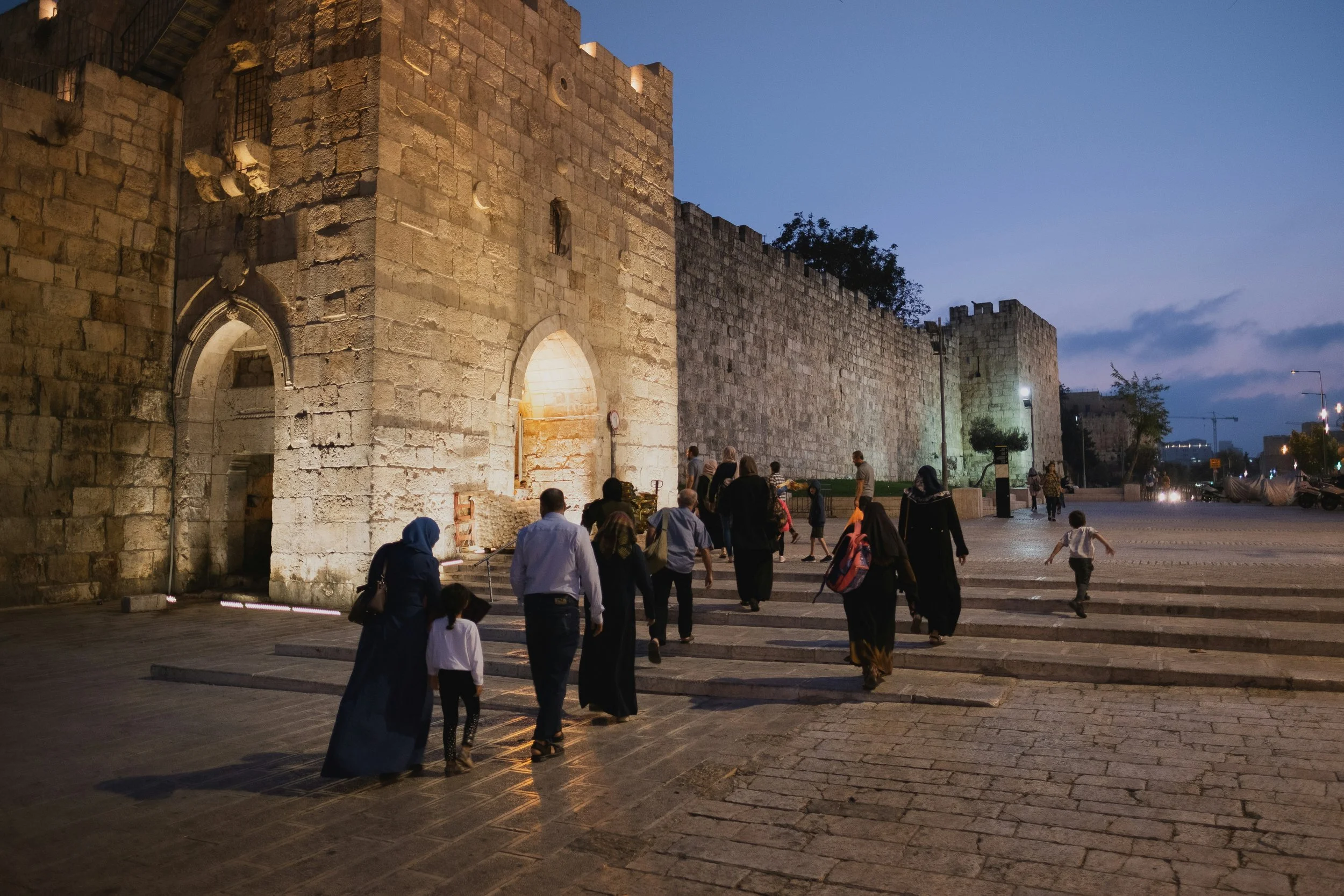 Jerusalem's ancient stone city walls illuminated by streetlights at dusk, with people walking in front, during the Holyland Horizons After Dark experience