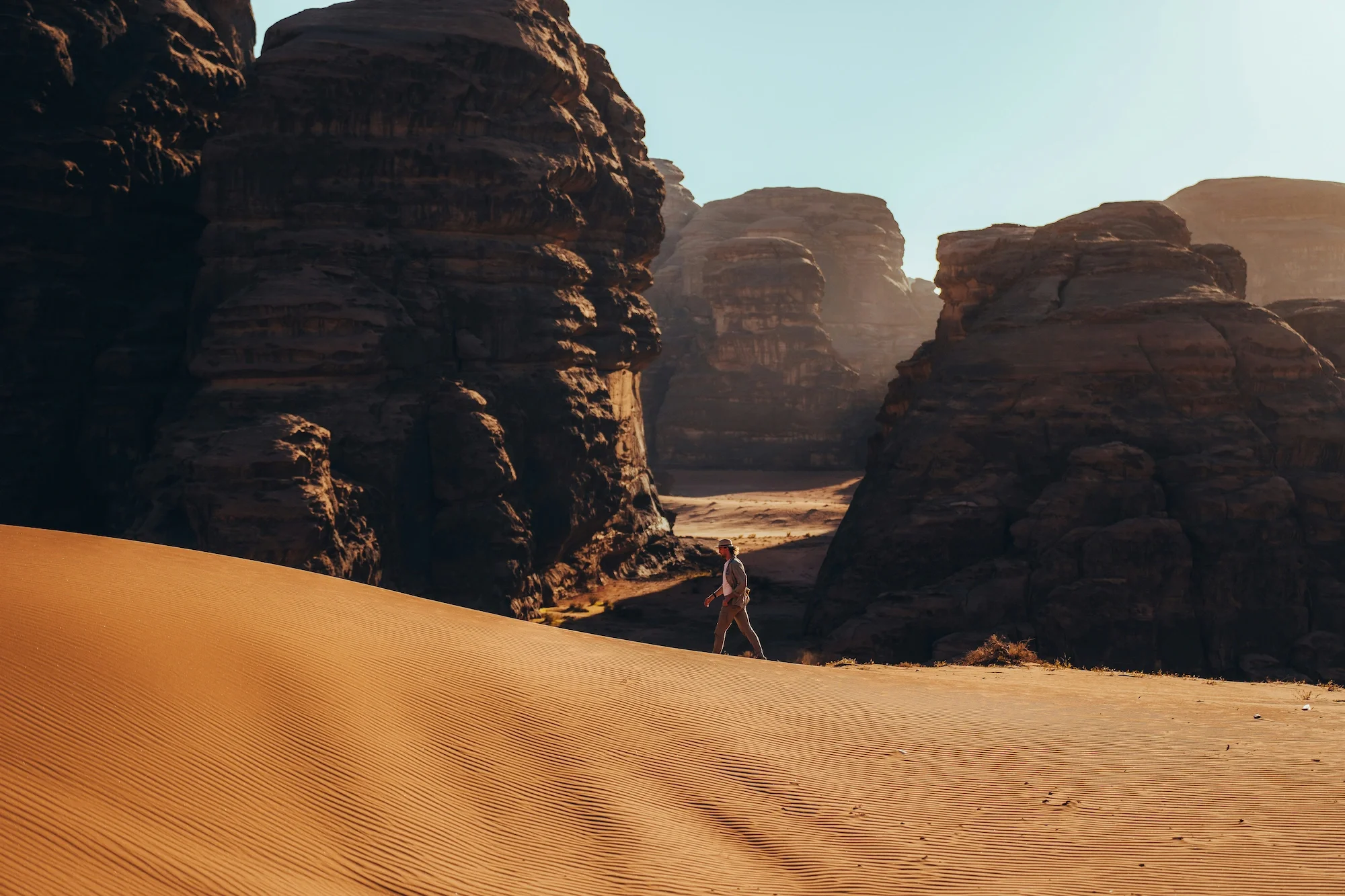 A person walking through a desert with large rock formations in the background and a clear sky.