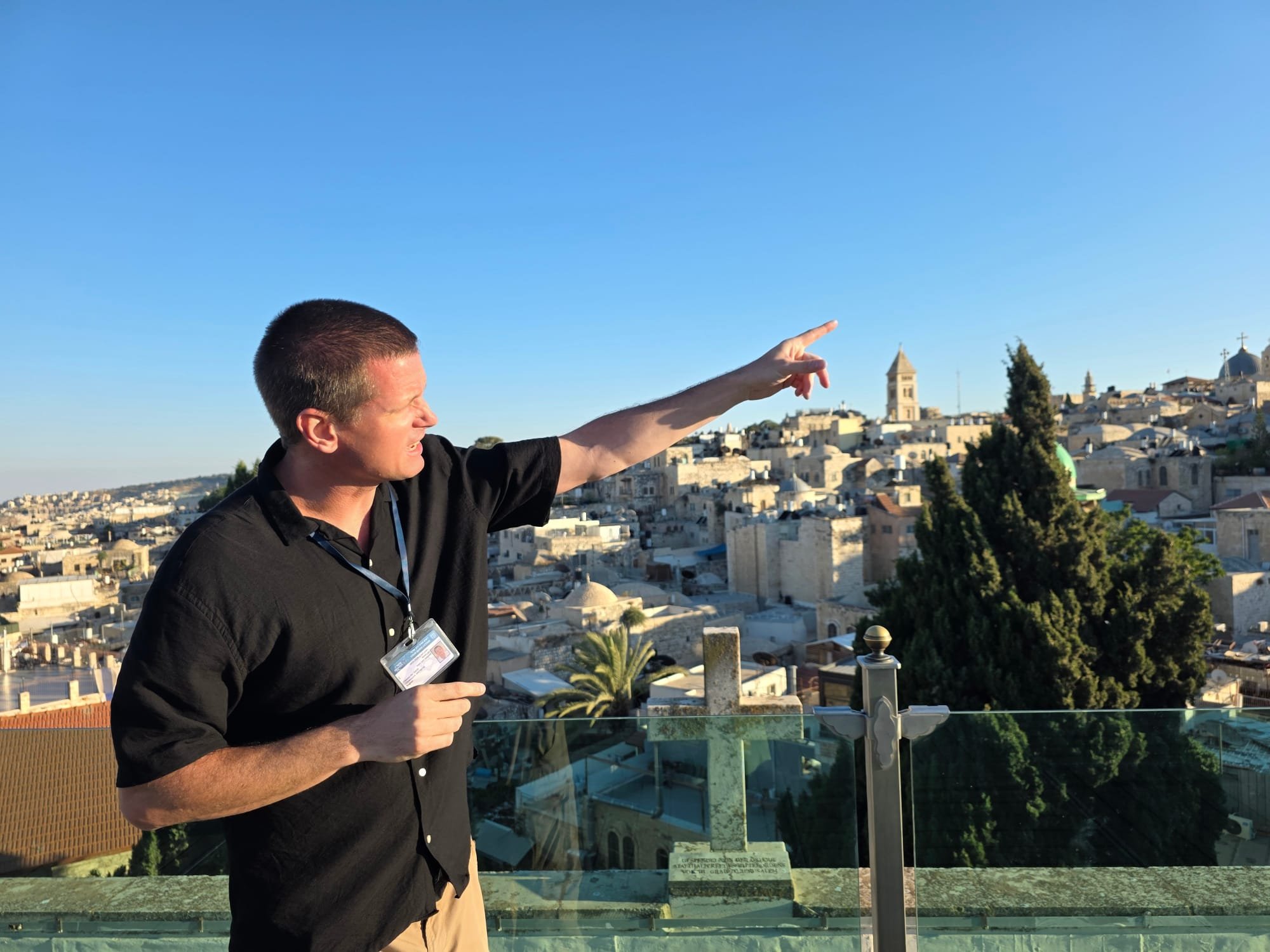 A man in a black shirt holding a visitor badge, pointing toward the distance with a cityscape in the background under a clear blue sky.