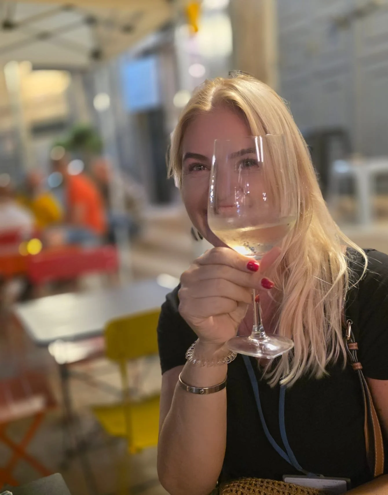 A woman with long blonde hair, wearing a black top and red nail polish, holding a glass of white wine while smiling at the camera in a restaurant or cafe setting.