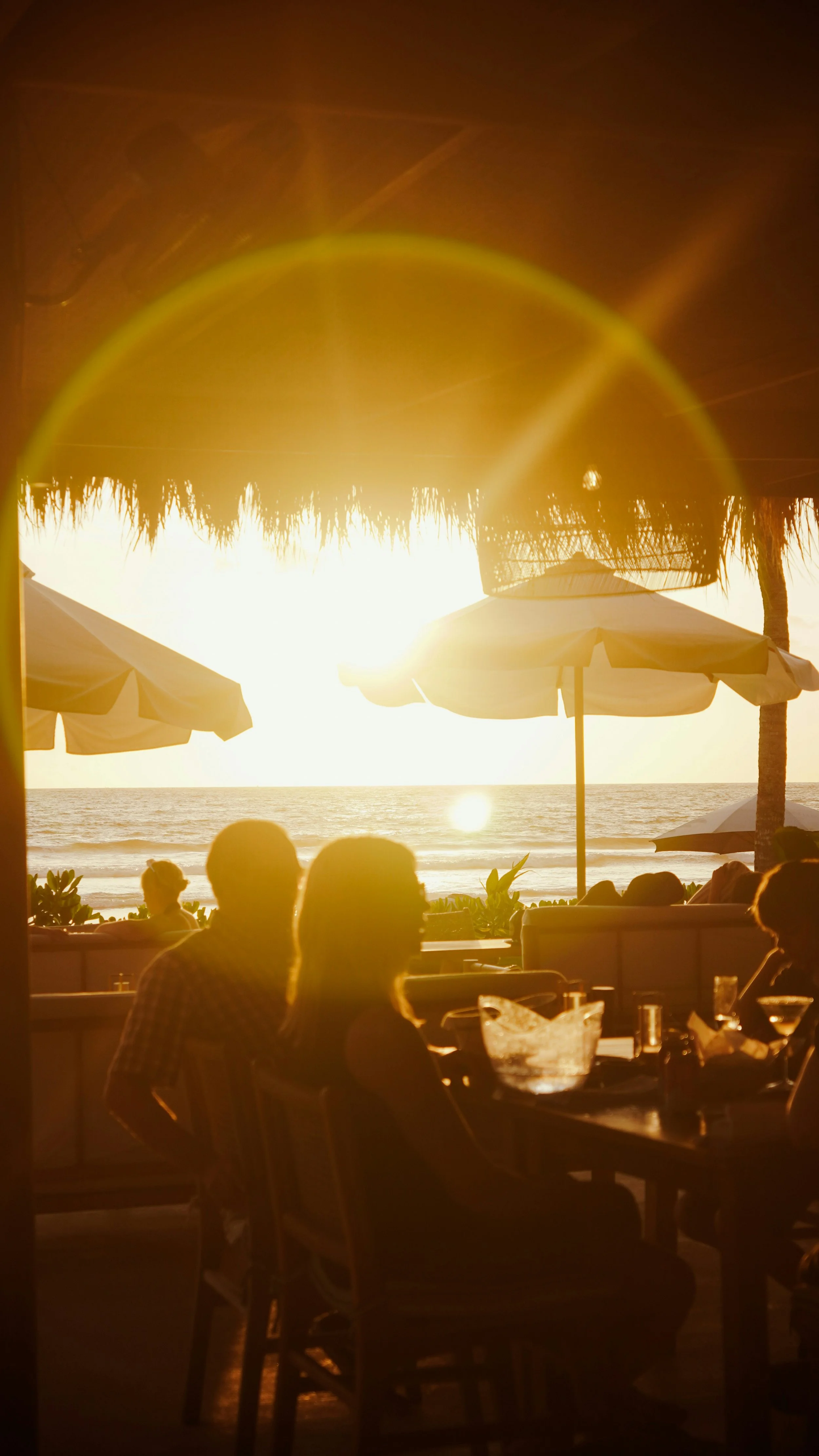 People sitting at a restaurant or bar with a view of the beach during sunset, with umbrellas and the ocean in the background.