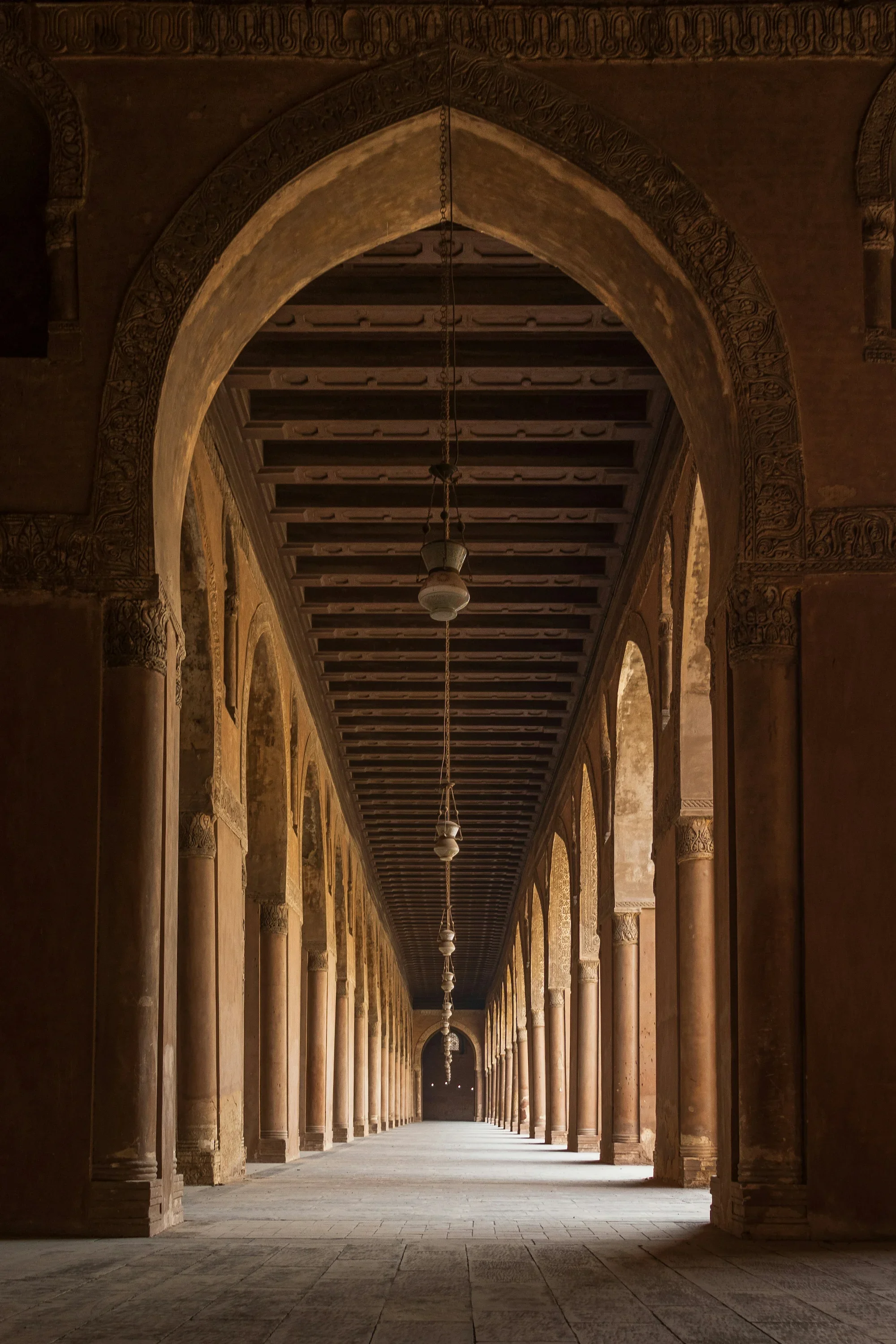 A long corridor with arches and columns on each side, with intricate carvings on the archways and a series of hanging lamps along the ceiling. The corridor appears historic and architectural, with sunlight illuminating the end of the hallway.