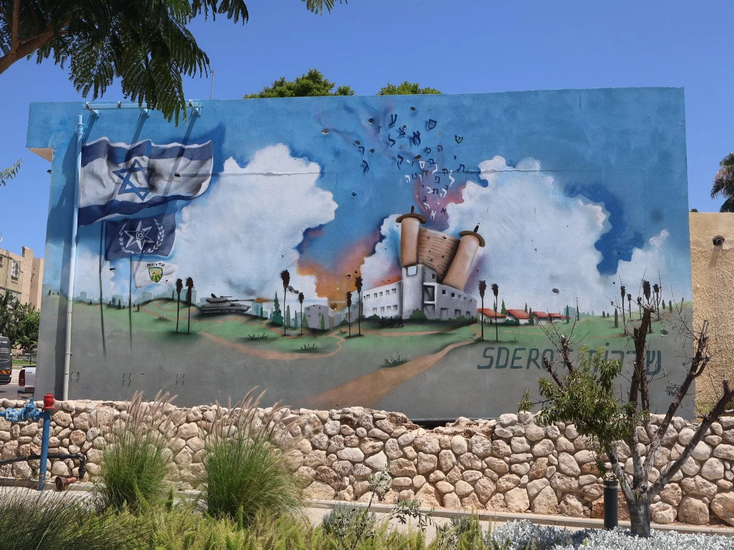 A mural depicting an industrial building with smoke billowing from chimneys, alongside flags including the Israeli flag. The scene includes trees and a grassy landscape with a clear blue sky.