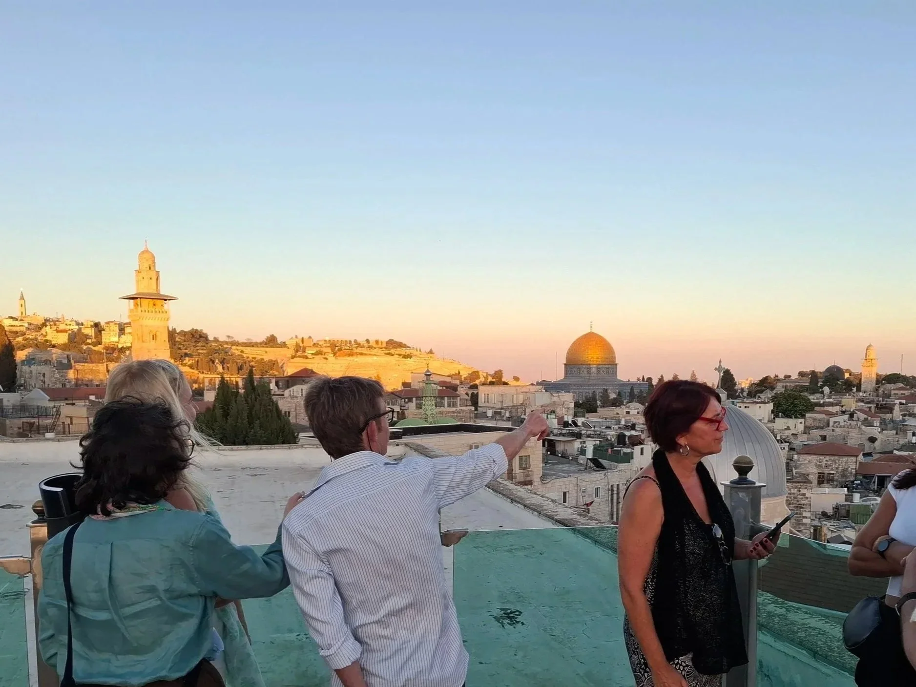 People standing on a rooftop observation deck with cityscape and famous landmarks in Jerusalem at sunset, including the Dome of the Rock and ancient towers.