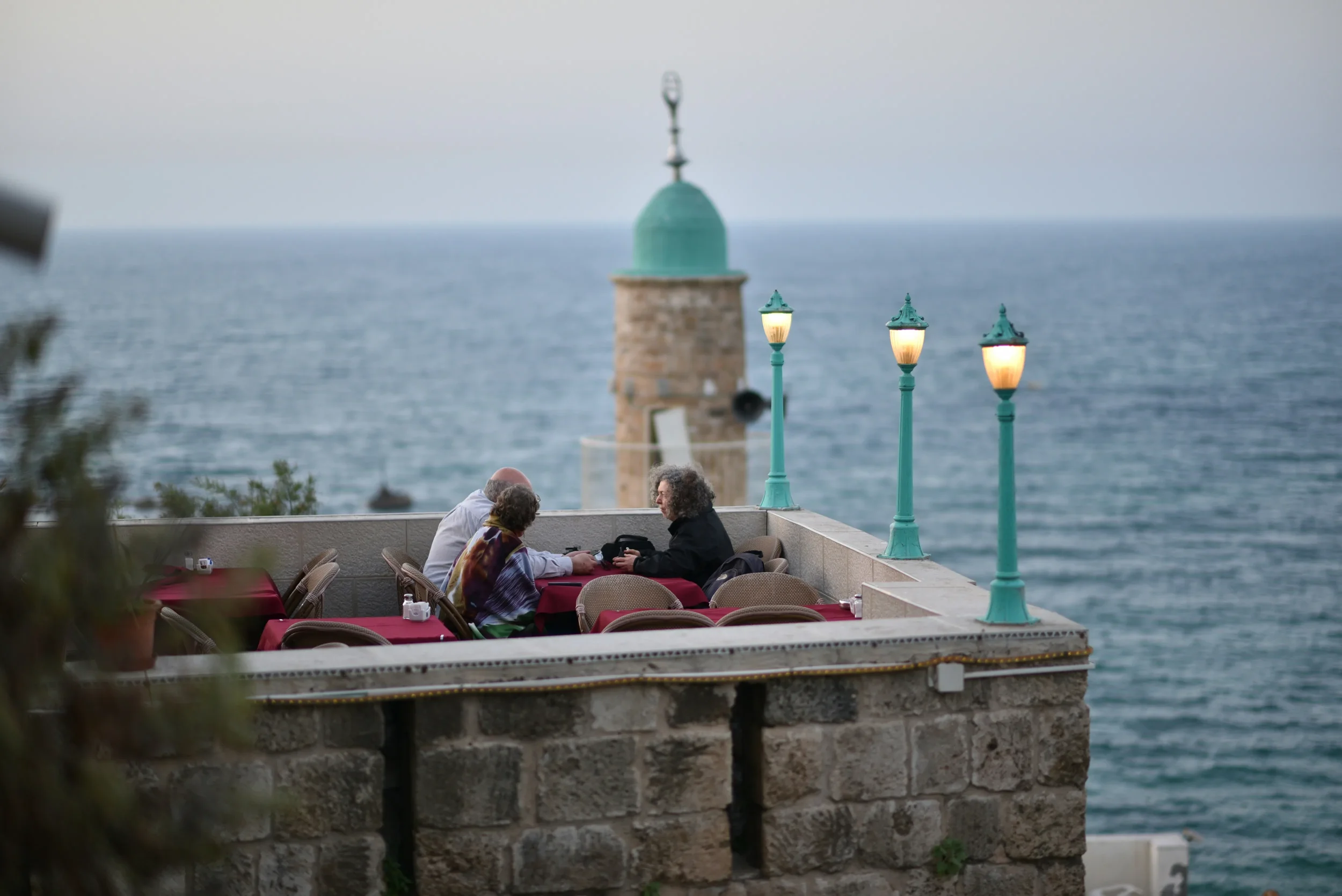 People sitting at tables on a terrace overlooking the ocean, with three vintage-style lampposts and a small tower with a green dome in the background.
