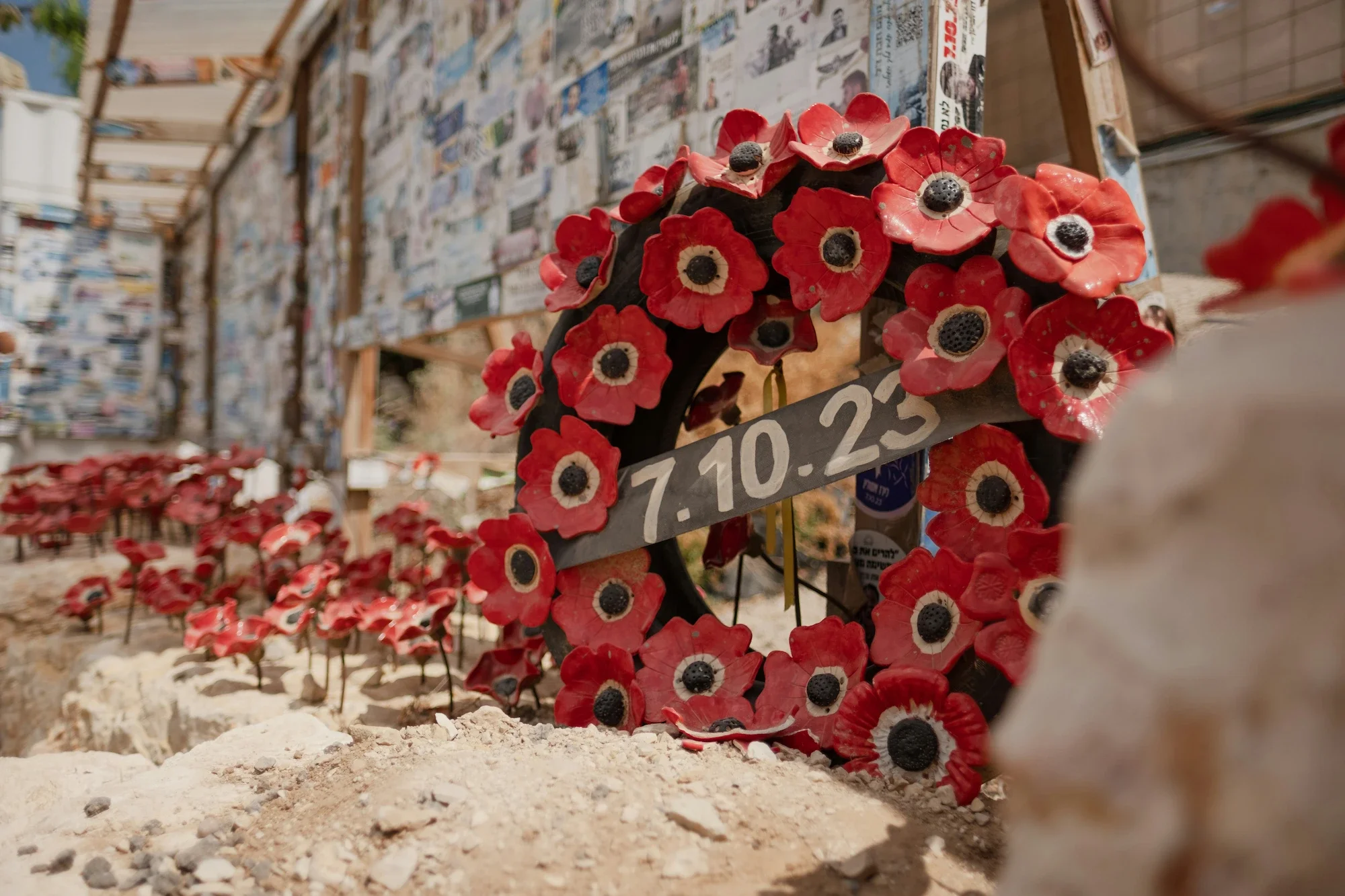 A wreath made of red ceramic poppies with black centers, sitting on a stone surface, with a blurred wall covered in photos or posters in the background.