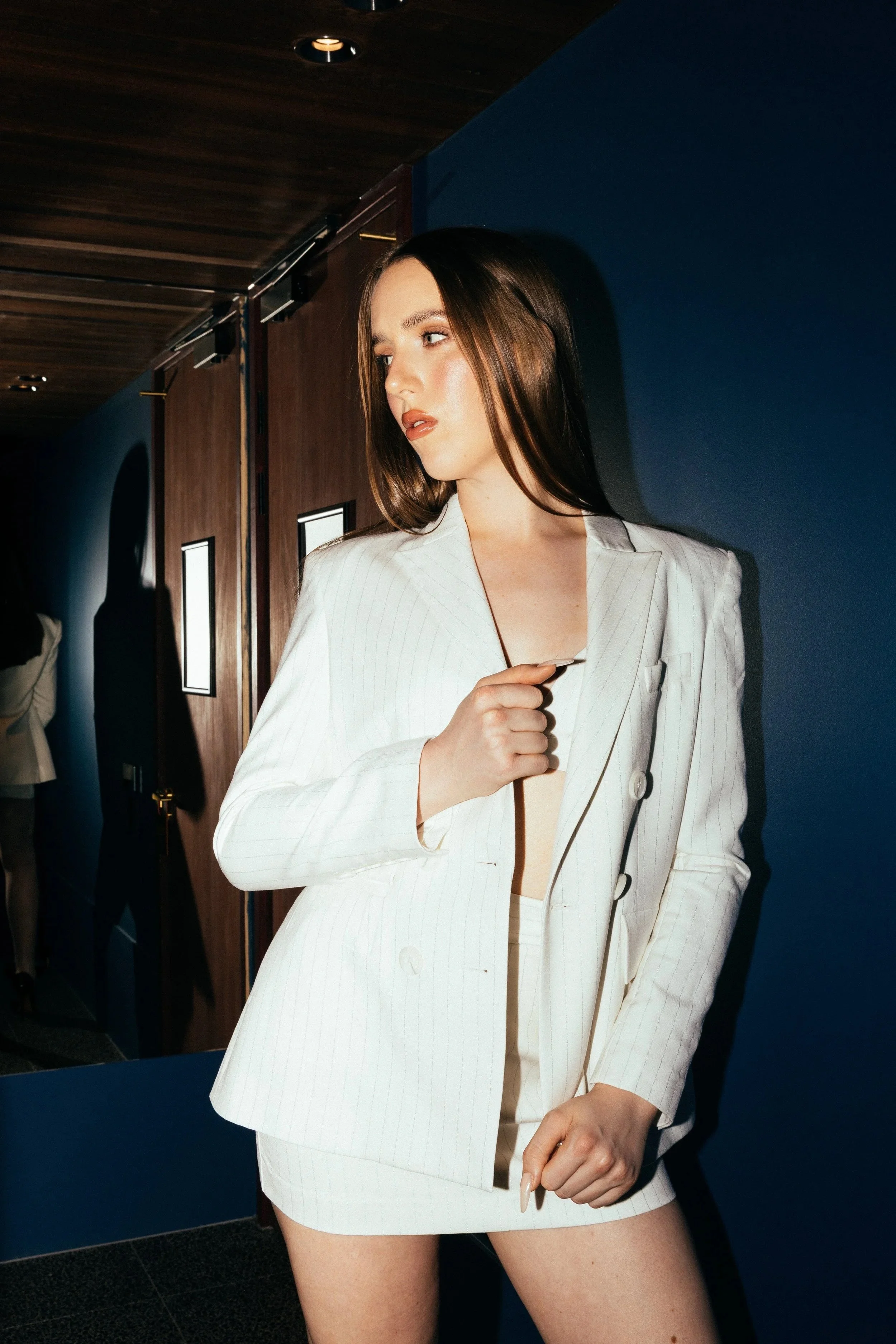 A woman with straight brown hair is wearing a white pinstripe blazer with a matching skirt. She is standing indoors against a dark blue wall, with hand on her chest and her head turned to the side.