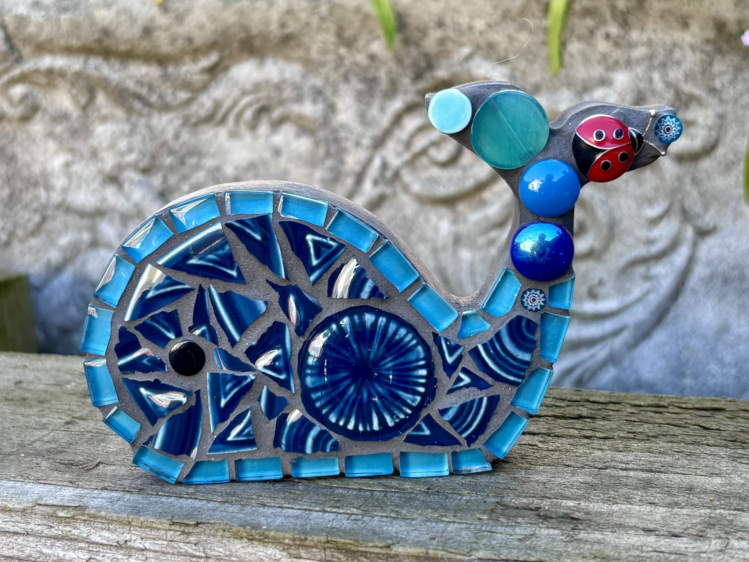 Colorful mosaic glass art piece with circular and geometric patterns in shades of blue, red, black, and teal, resting on a weathered wooden surface outdoors.
