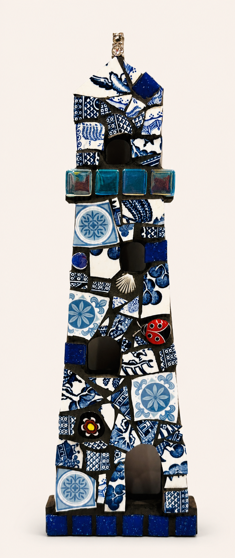 Tall ceramic sculpture resembling a stylized tower or house, made of irregularly shaped blue and white tiles with various patterns, including floral and geometric designs, decorated with small glass blocks, a ladybug figurine, and other decorative elements.