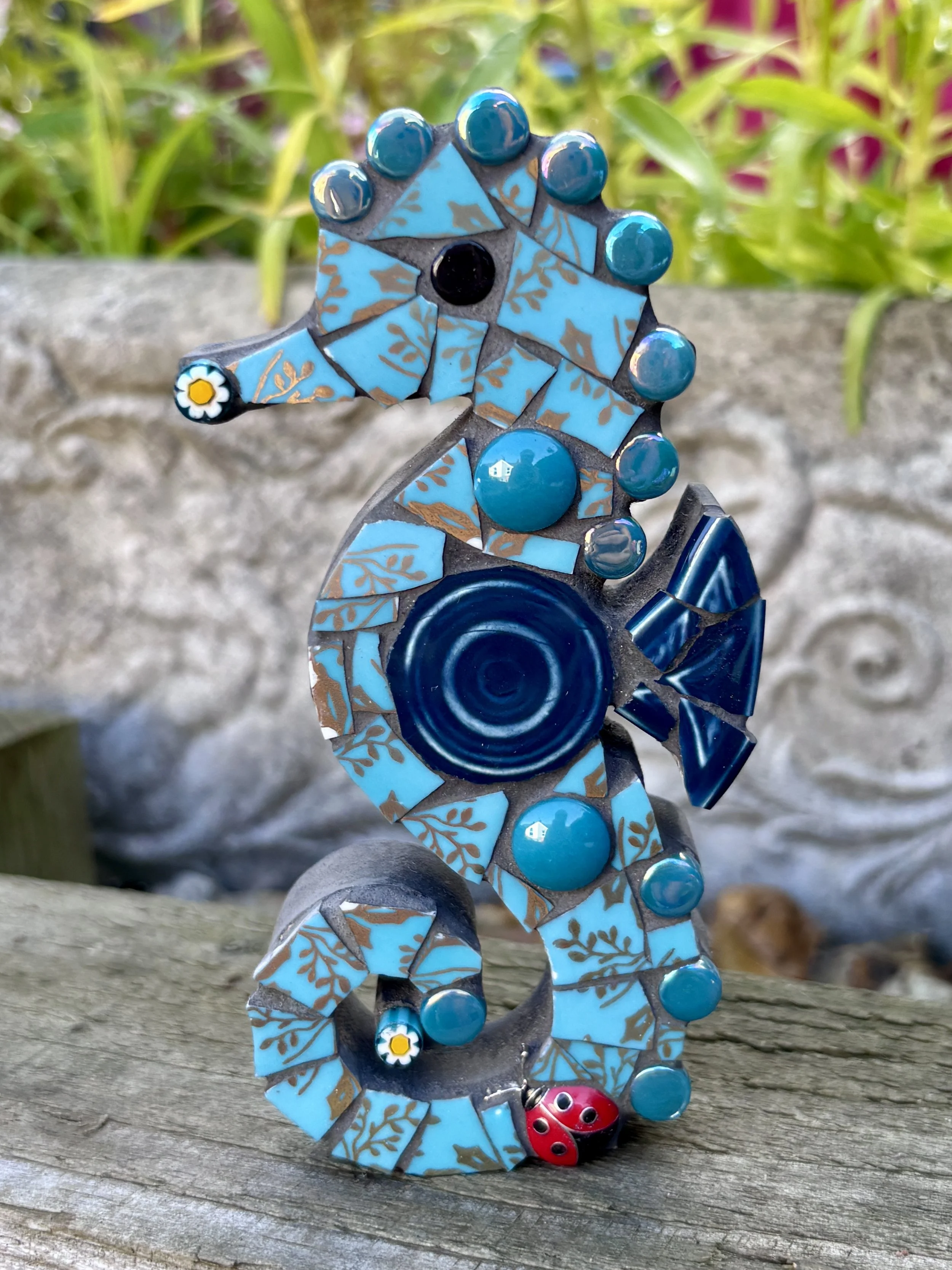 A mosaic seahorse sculpture with blue and brown tile patterns, topped with turquoise beads, a white daisy flower on tail, and a red ladybug at the base, set on a weathered wooden surface with green plants in the background.