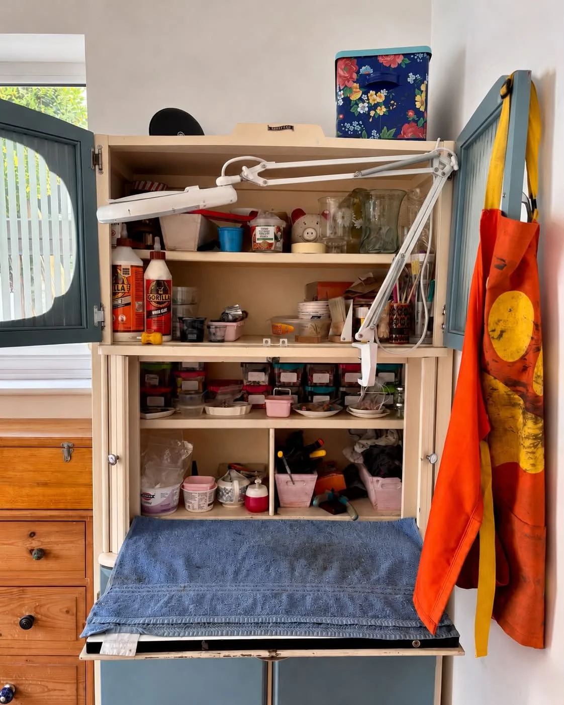 A tabletop craft station with shelves filled with art supplies, bottles of glue, jars, containers, and various crafting tools, with an orange apron hanging on the side and a blue towel draped over the surface.