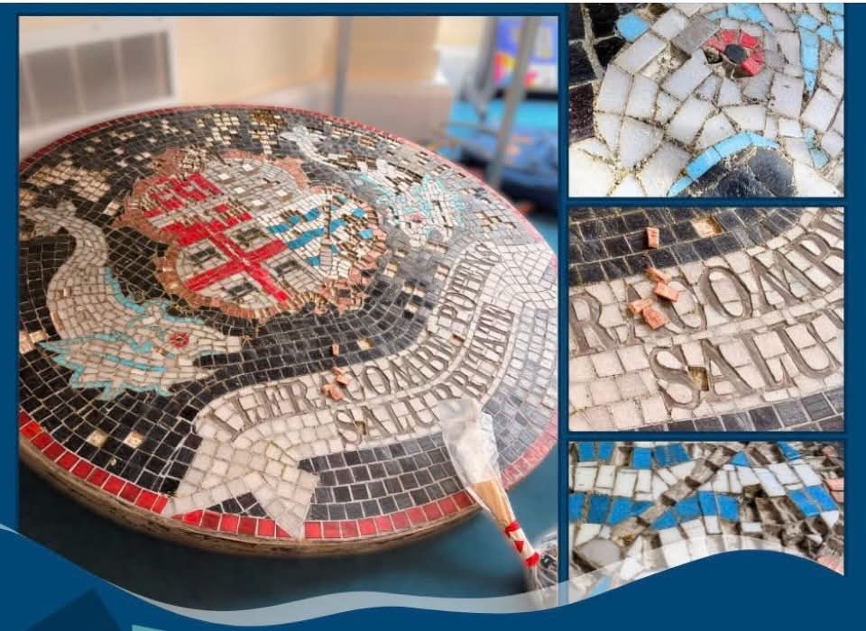 Close-up of a mosaic art piece with a shield emblem, wings, and Cyrillic text, showing damaged tiles and debris, with a blue and red background.