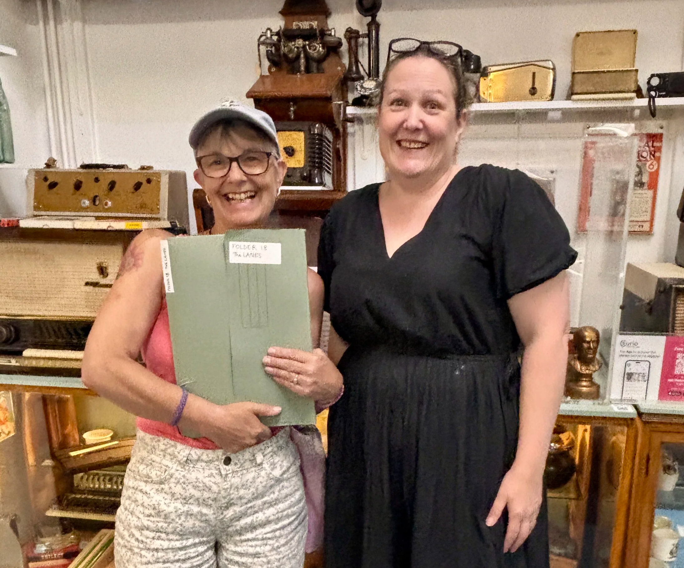 Two women smiling and standing close together in a room with various antique items and shelves in the background. One woman is holding a green book.