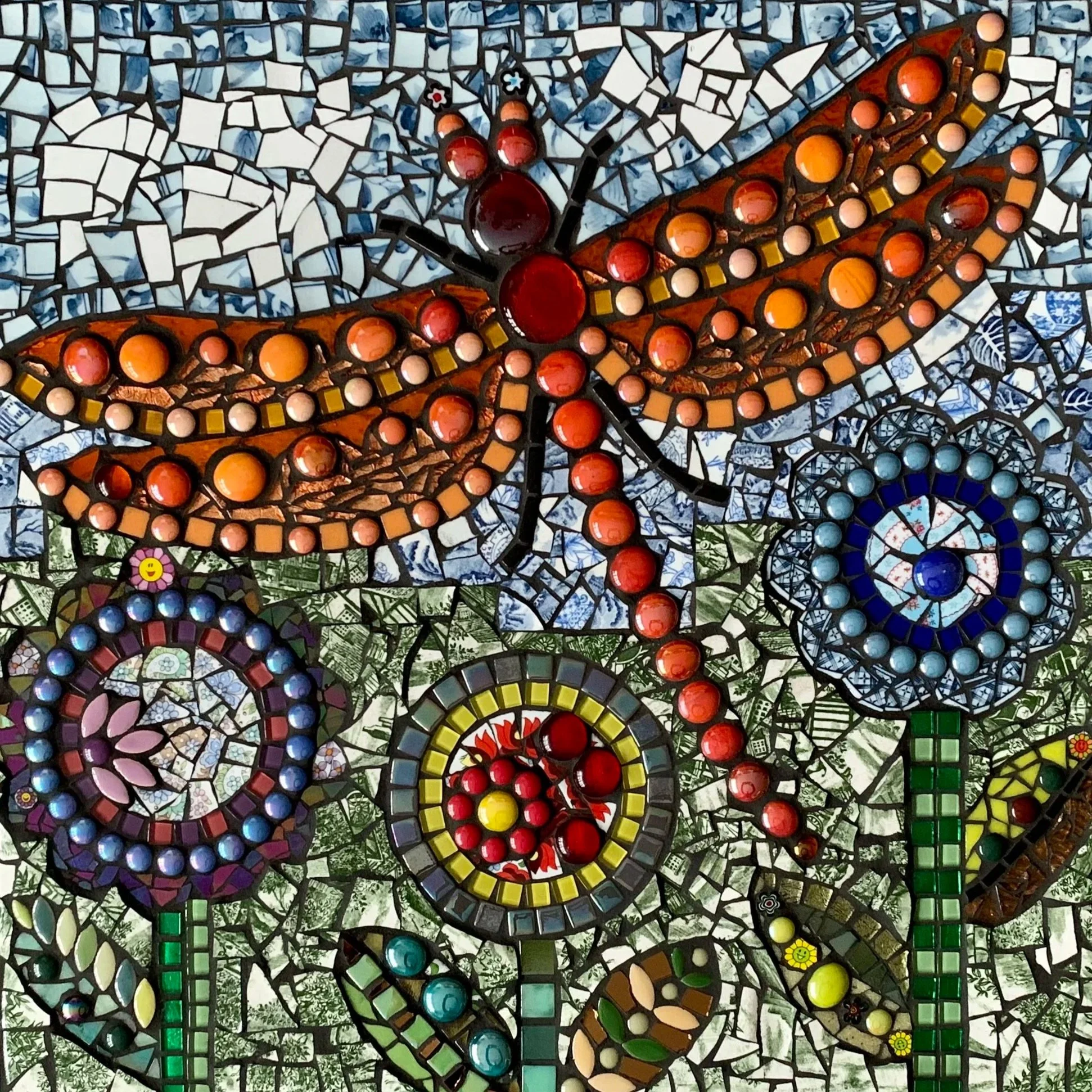 Colorful mosaic artwork featuring a dragonfly design with red, orange, and blue circular tiles forming the body and wings, set against a background of small, irregularly shaped tiles in shades of white, gray, and green.