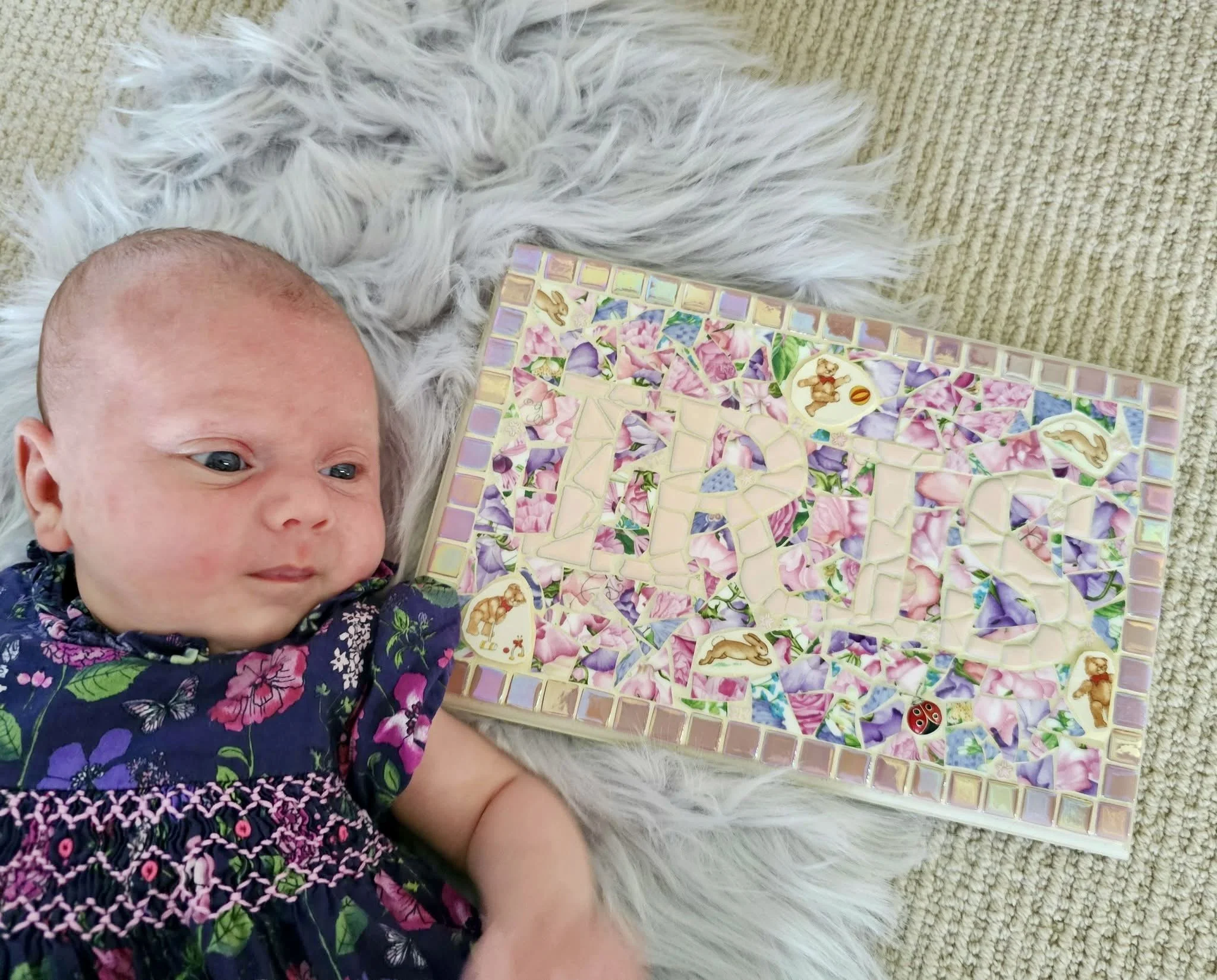A baby with blue eyes and fair skin lying on a fluffy white blanket next to a colorful mosaic tile game, with a small ladybug on the tiles.