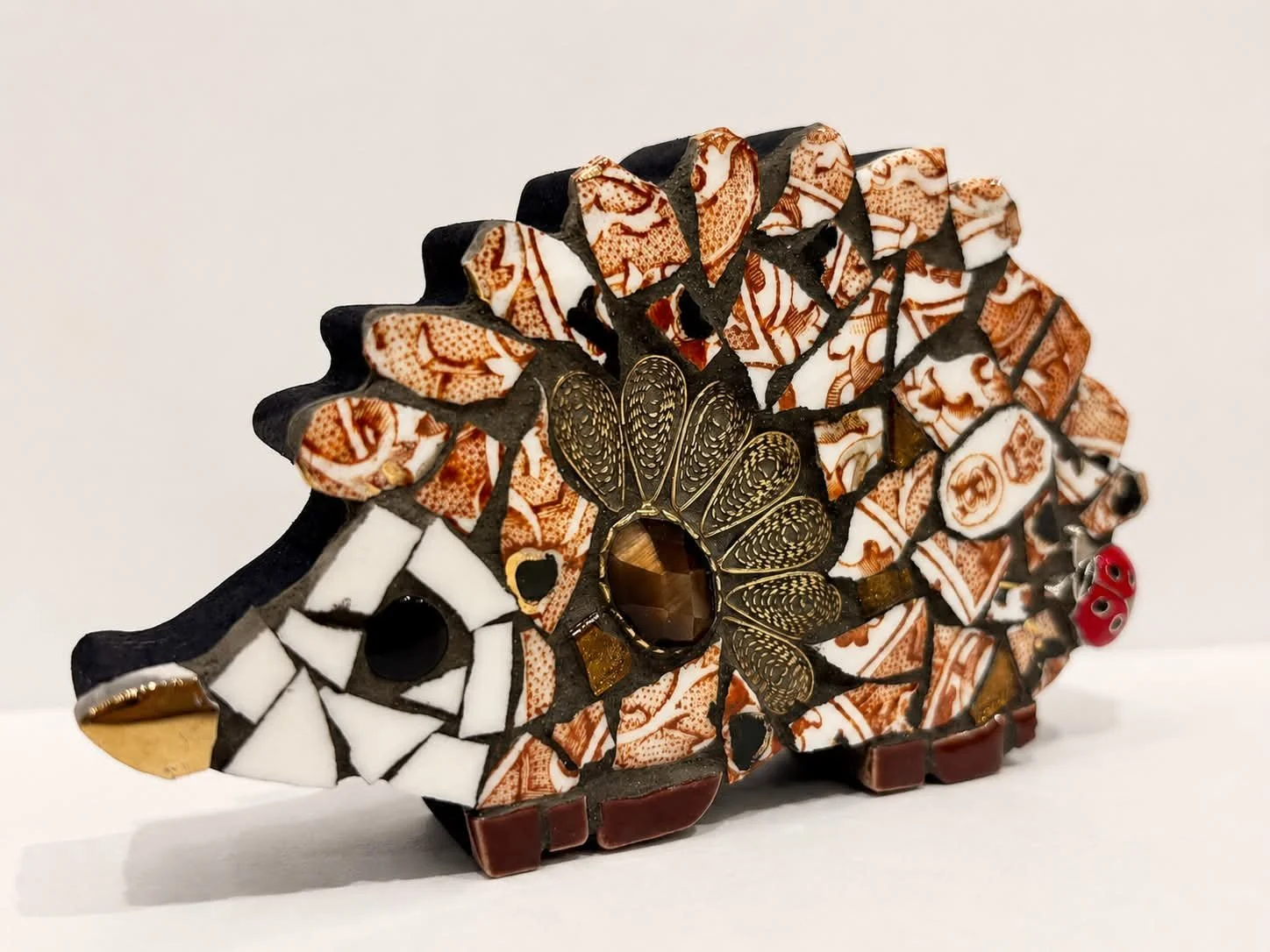 A colorful mosaic sculpture of a hedgehog made of various patterned and textured materials, including glass, beads, and metal. The sculpture's body features intricate designs and a large central gemstone, with small decorative elements like beads and metallic accents.