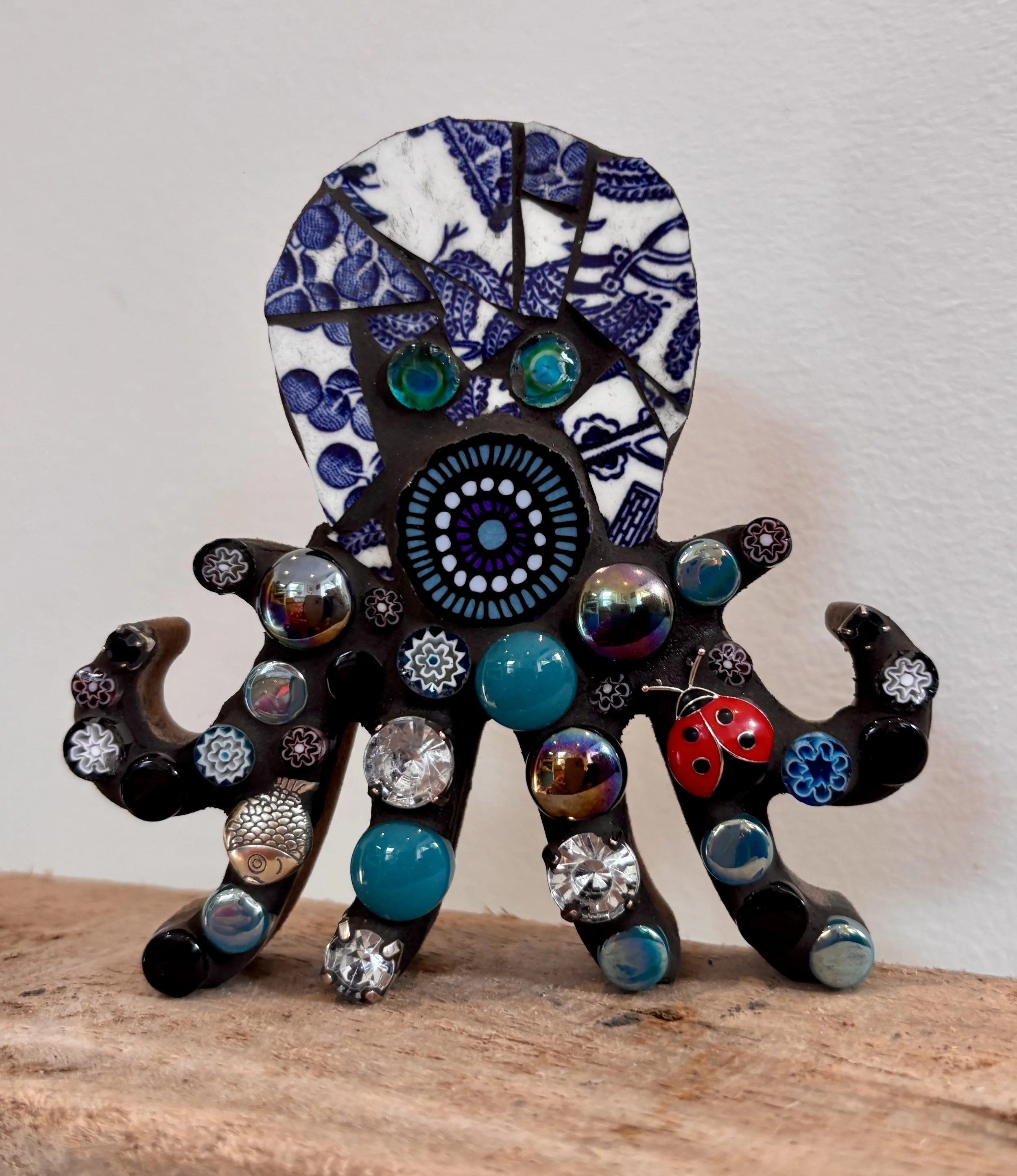 A decorative zebra figurine made of black, white, and blue ceramic pieces, featuring colorful glass and metal beads, standing on a wooden surface against a plain wall background.