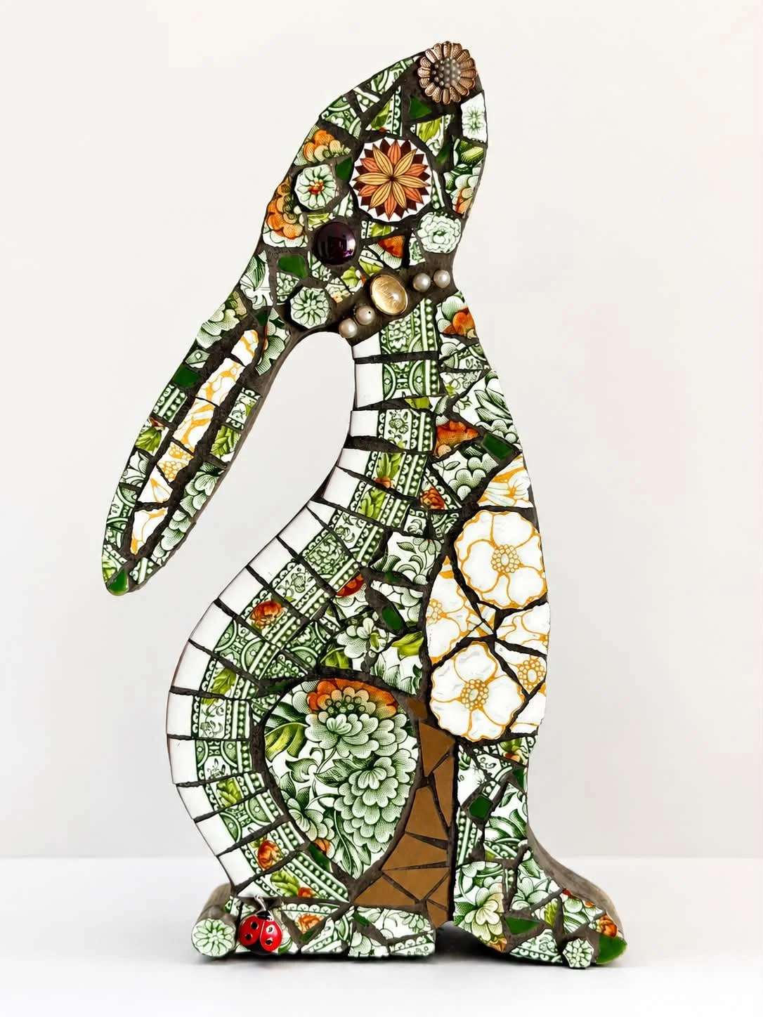 Decorative mosaic rabbit sculpture with floral and geometric patterns, adorned with various pins and buttons, against a white background.