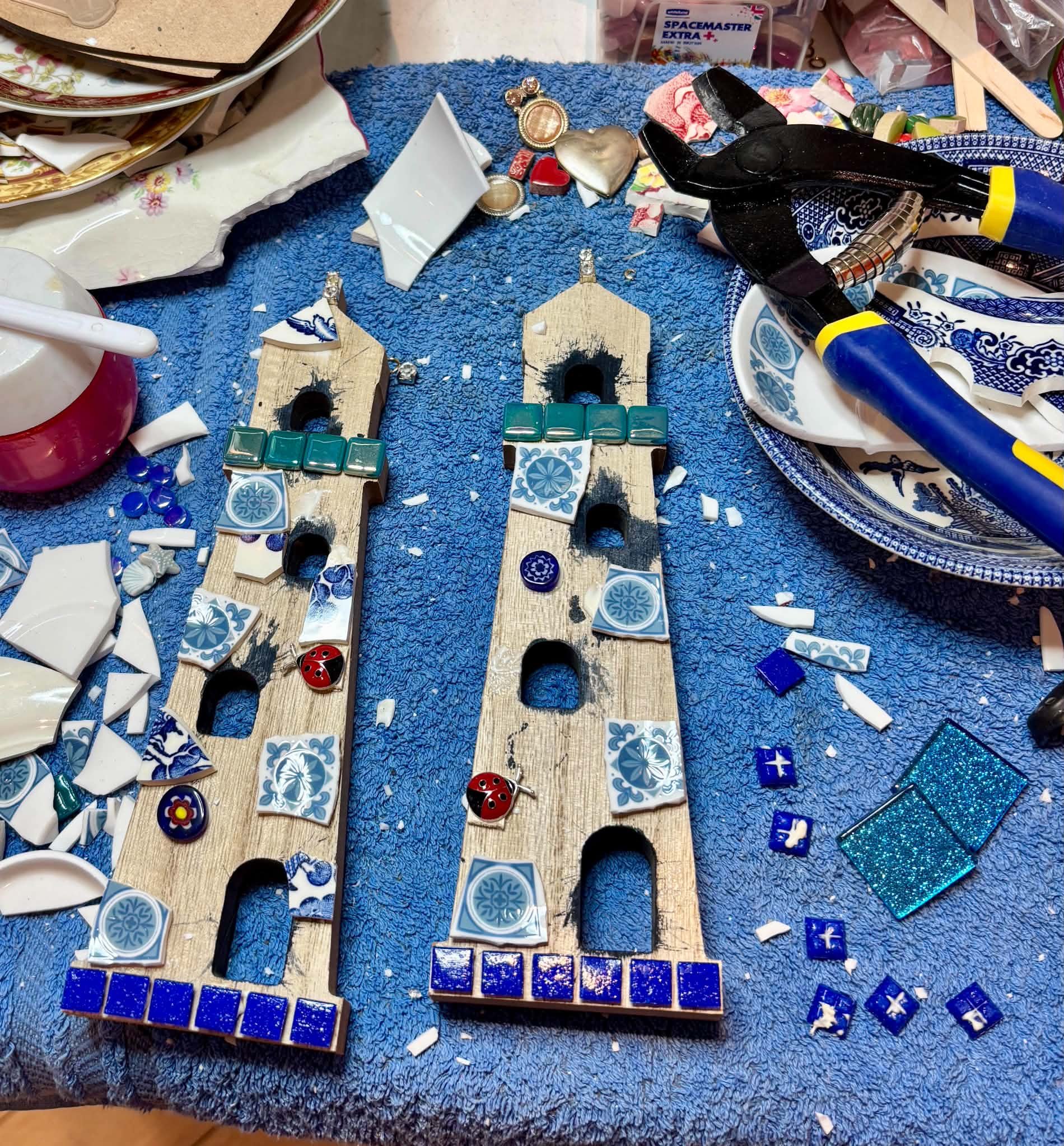 Close-up of a cluttered tabletop with broken blue and white patterned tiles, small game pieces, a black and yellow glue gun, jewelry, and papers on a blue fabric surface.