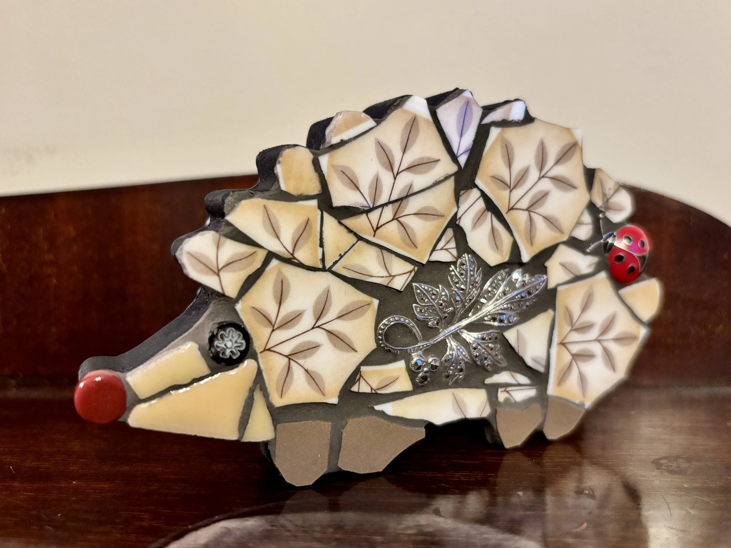 mosaic hedgehog made of cream, black, and purple stones with tan leaf patterns, red bead accents, and silver floral embellishments on a wooden surface.