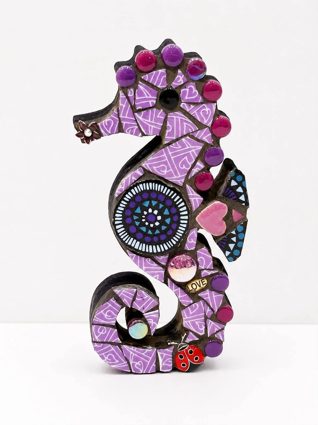 Decorative metal seahorse sculpture with purple, pink, and black accents, featuring various charms and patterns, set against a plain white background.