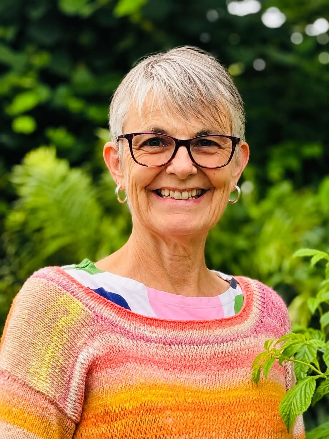 A smiling elderly woman with glasses and short gray hair standing outdoors with green foliage in the background.