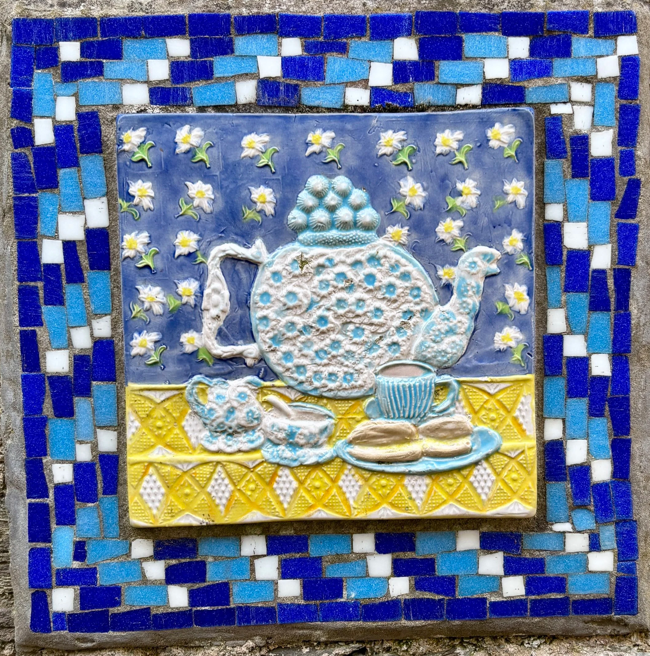Mosaic tile artwork with a blue border and design consisting of white flowers with yellow centers, green leaves, and various patterned tiles in blue, yellow, and white.