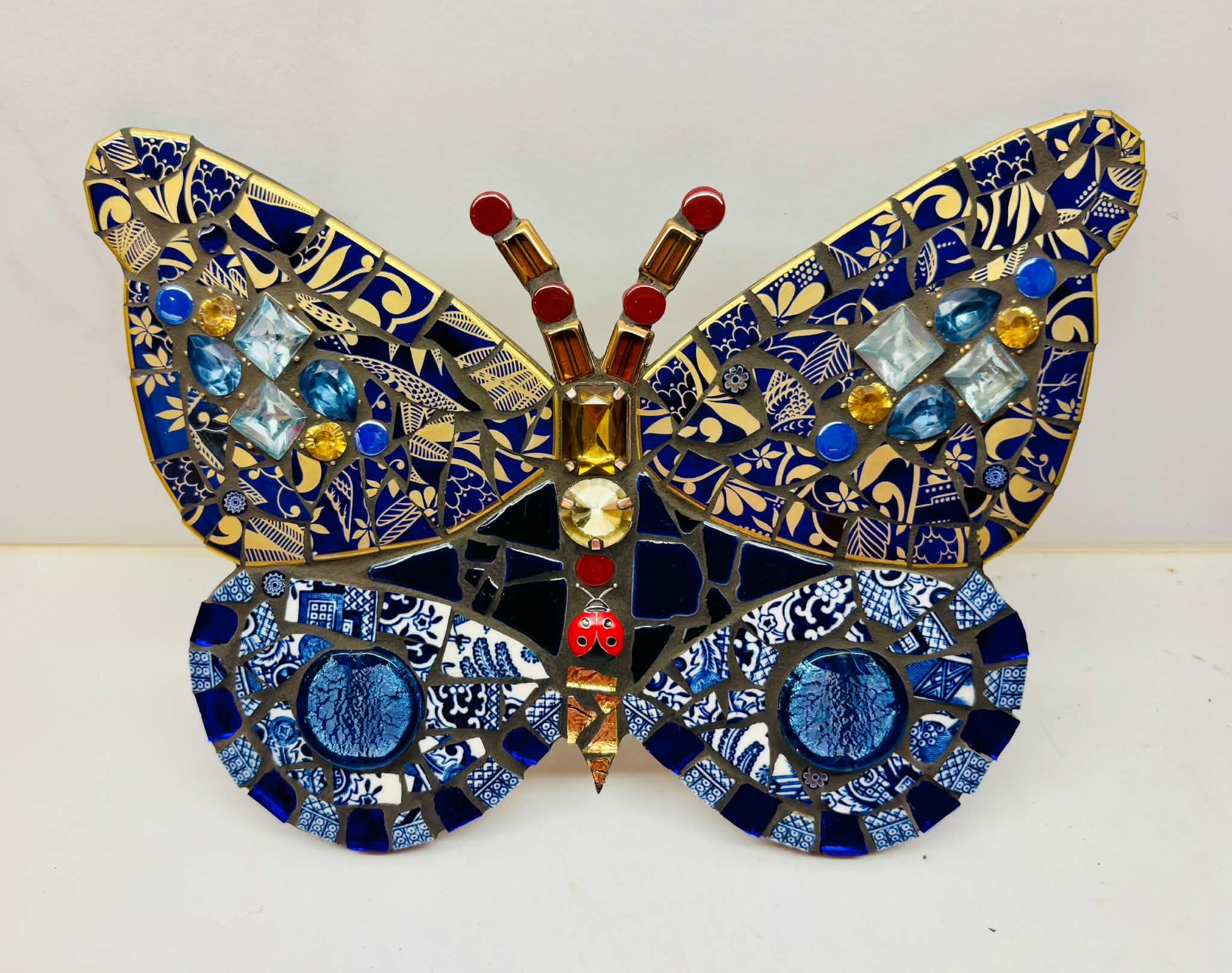 Decorative butterfly made of blue and gold patterned ceramic or glass pieces with embedded colorful gems, set against a plain white background.