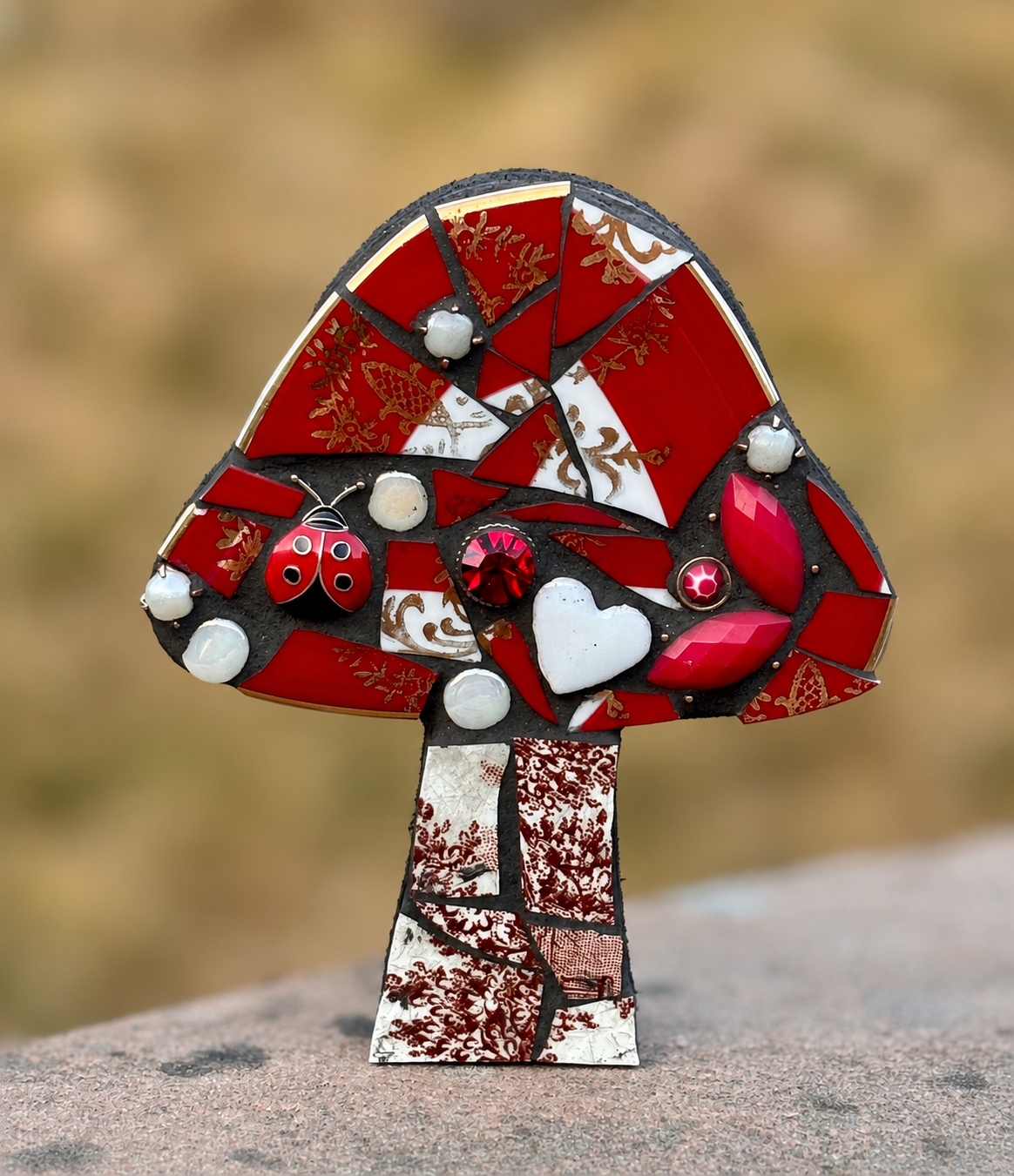 A colorful mosaic art piece of a mushroom with a red cap featuring gold patterns, decorated with various embellishments including a red ladybug, a white heart, red and white stones, and white pearls on a blurred outdoor background.