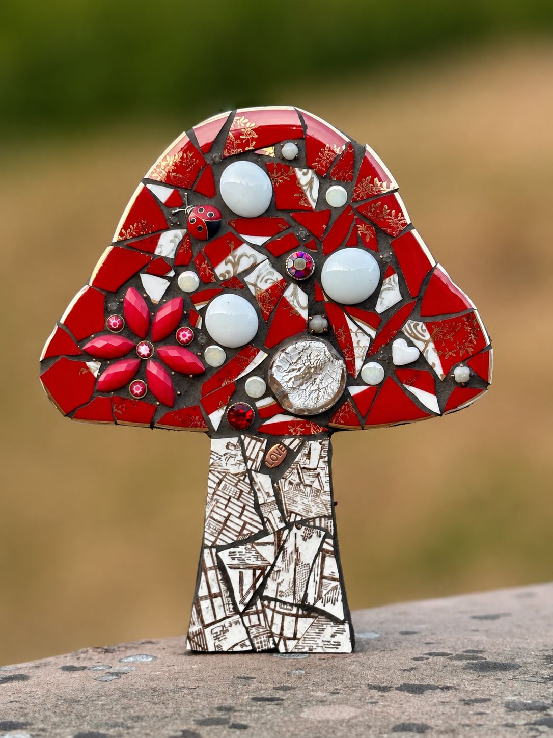 Mosaic art of a red, white, and black mushroom with floral and abstract patterns, standing on a textured surface outdoors.