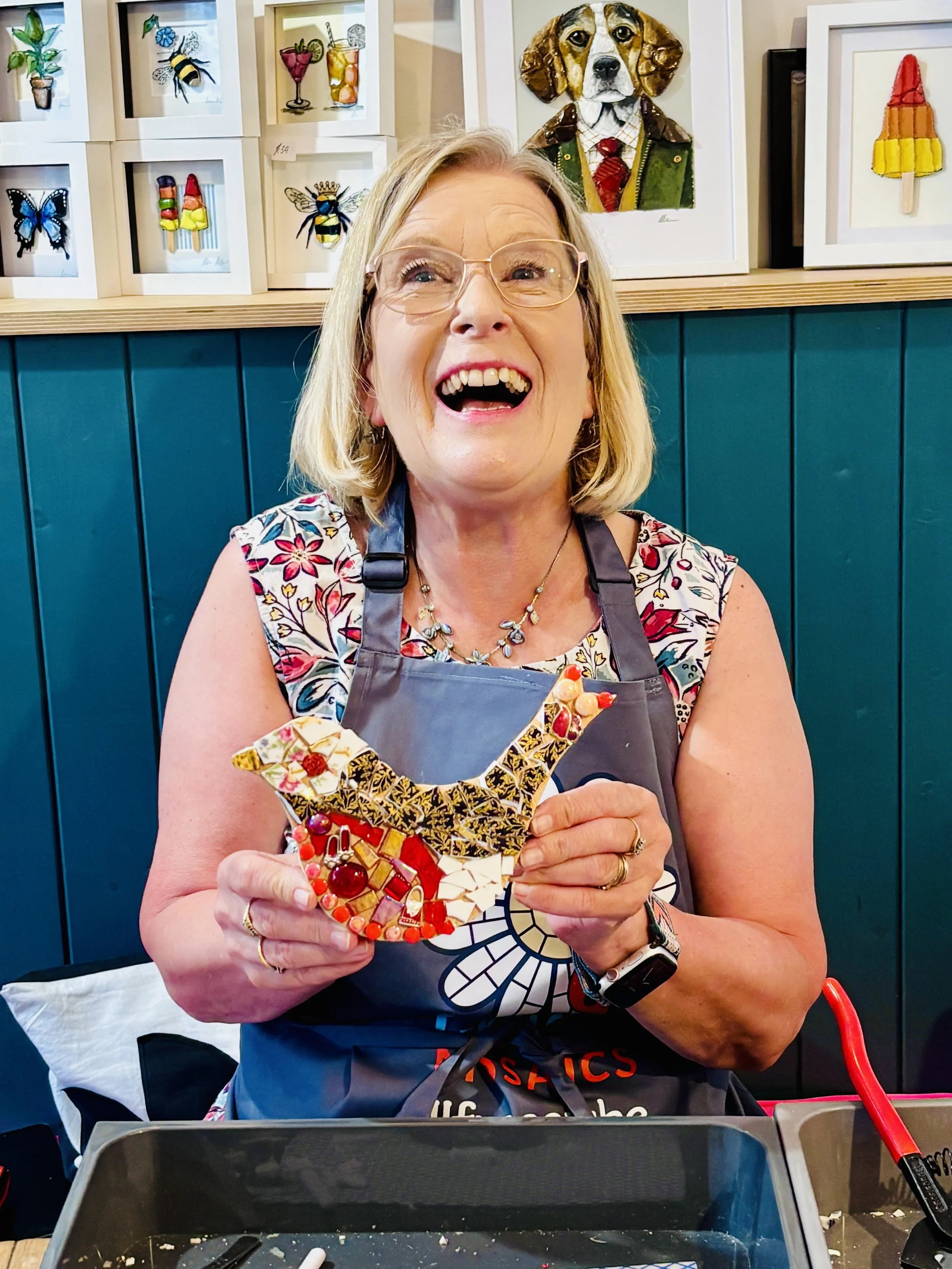A woman with blonde hair, glasses, and colorful floral top smiling widely while holding a decorated heart-shaped mosaic art piece. She is wearing a gray apron and a wristwatch. Behind her is a teal wall with framed artwork of insects, animals, and candy-themed objects, and various art supplies and tools are on the table in front of her.
