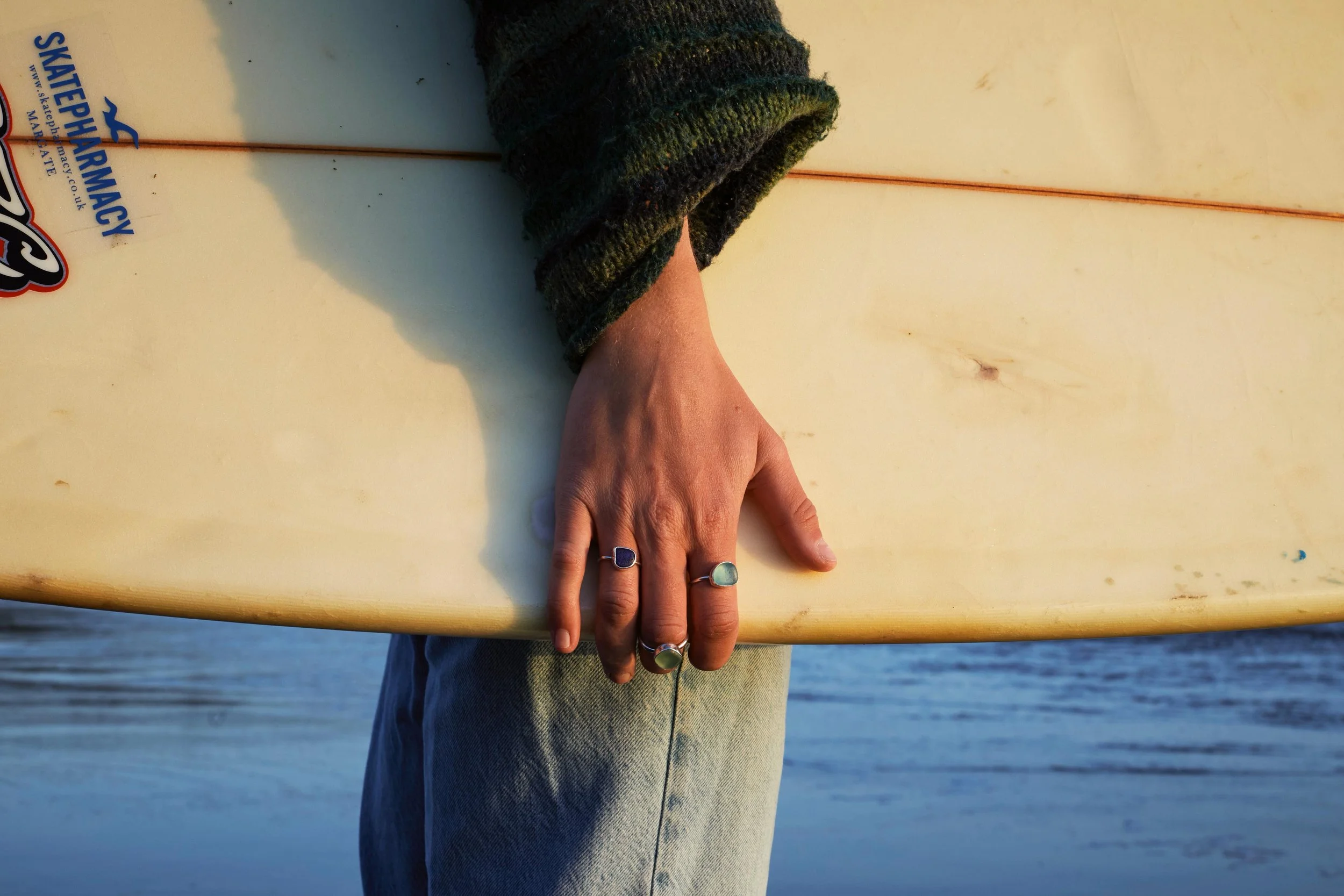 Hand with rings resting on a surfboard near the beach, wearing a sweater with a background of ocean and sky.