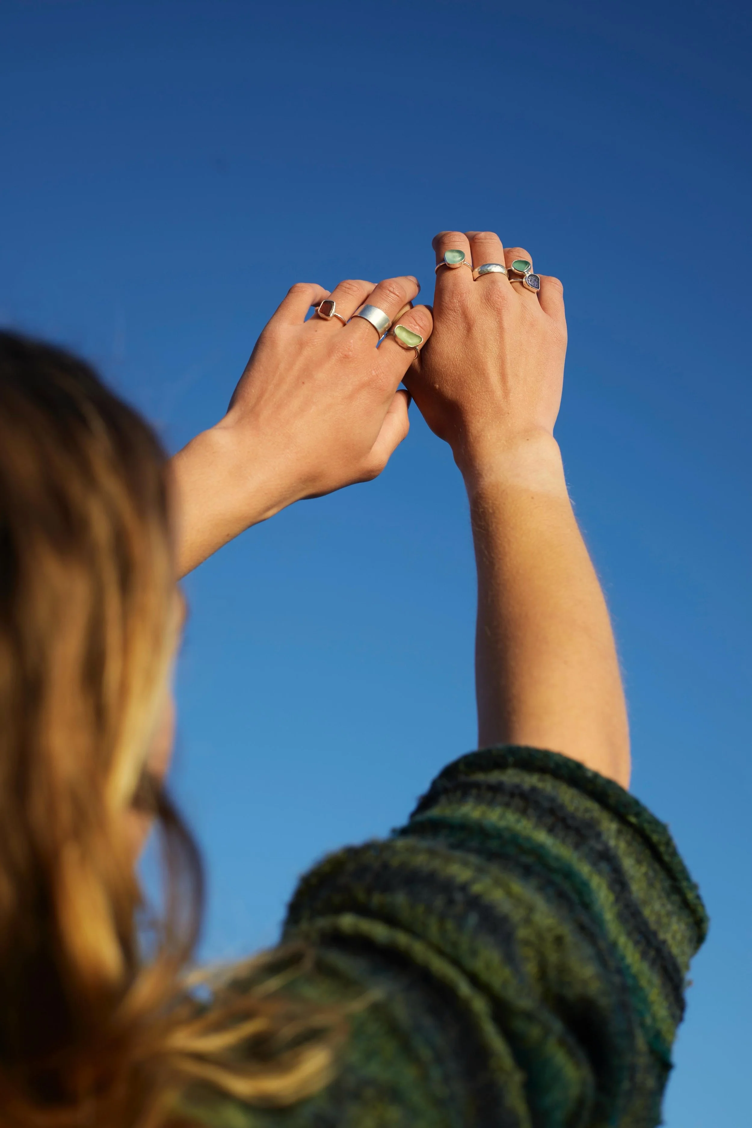 Two hands with multiple silver rings, held against a clear blue sky. One hand is also wearing a green ring or gemstone. Part of a person's head with curly hair and a striped green and black long-sleeve shirt is visible in the foreground.