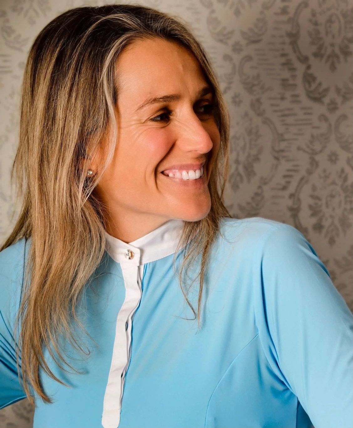 Donna che sorride indossando abbigliamento sportivo equestre, capelli castani e sfondo decorato.