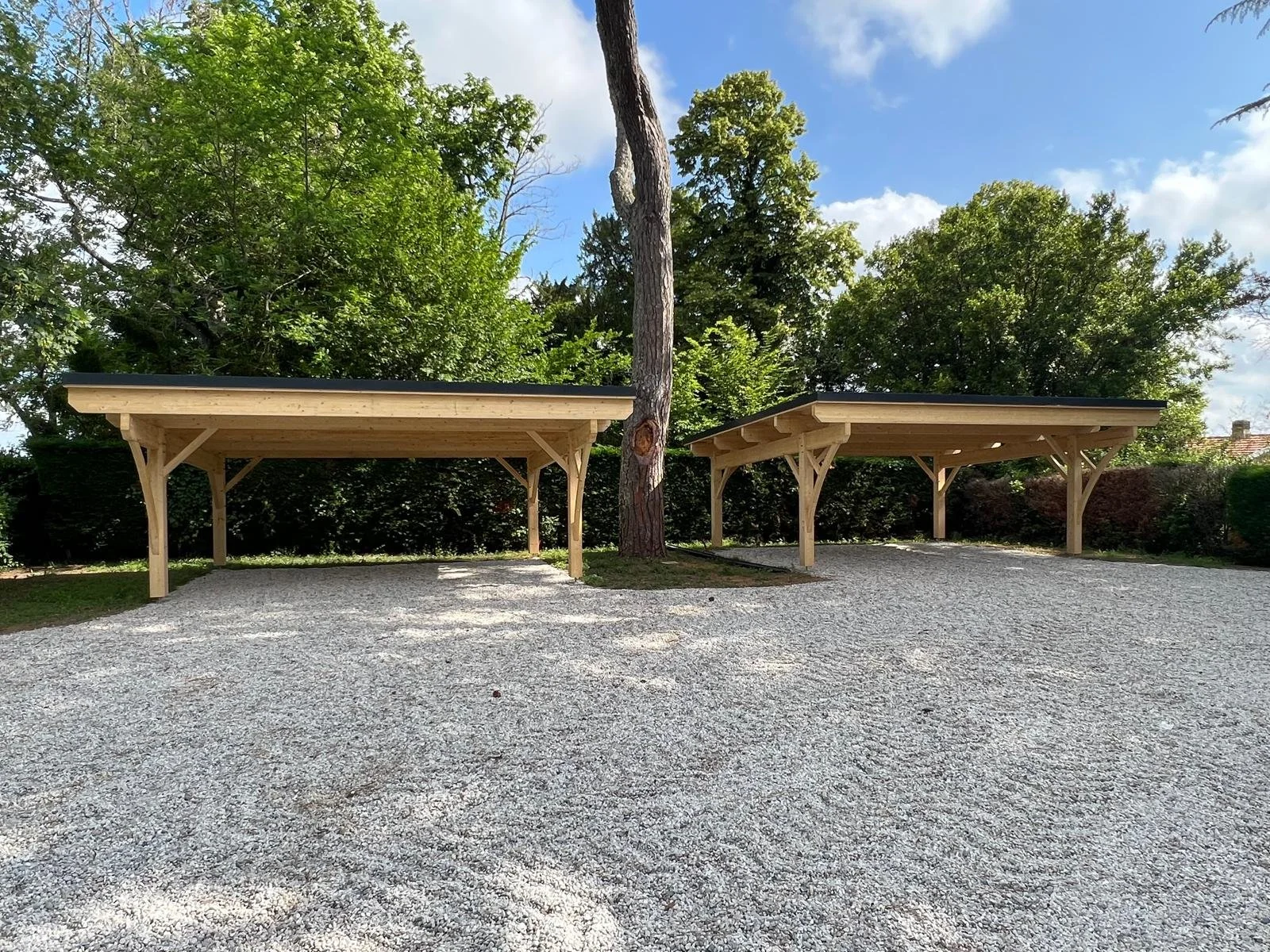 Deux abris en bois avec toit noir, situés dans un espace en gravier, entourés d'arbres verts et un ciel bleu avec quelques nuages.