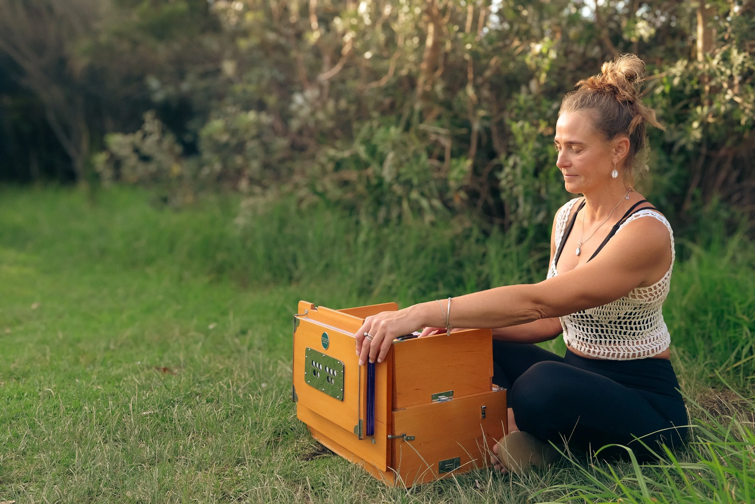 Yoga teacher Narelle with hair tied up in a bun, wearing a crochet top and jewellery, sitting outdoors on grass, playing a wooden harmonium in front of her.