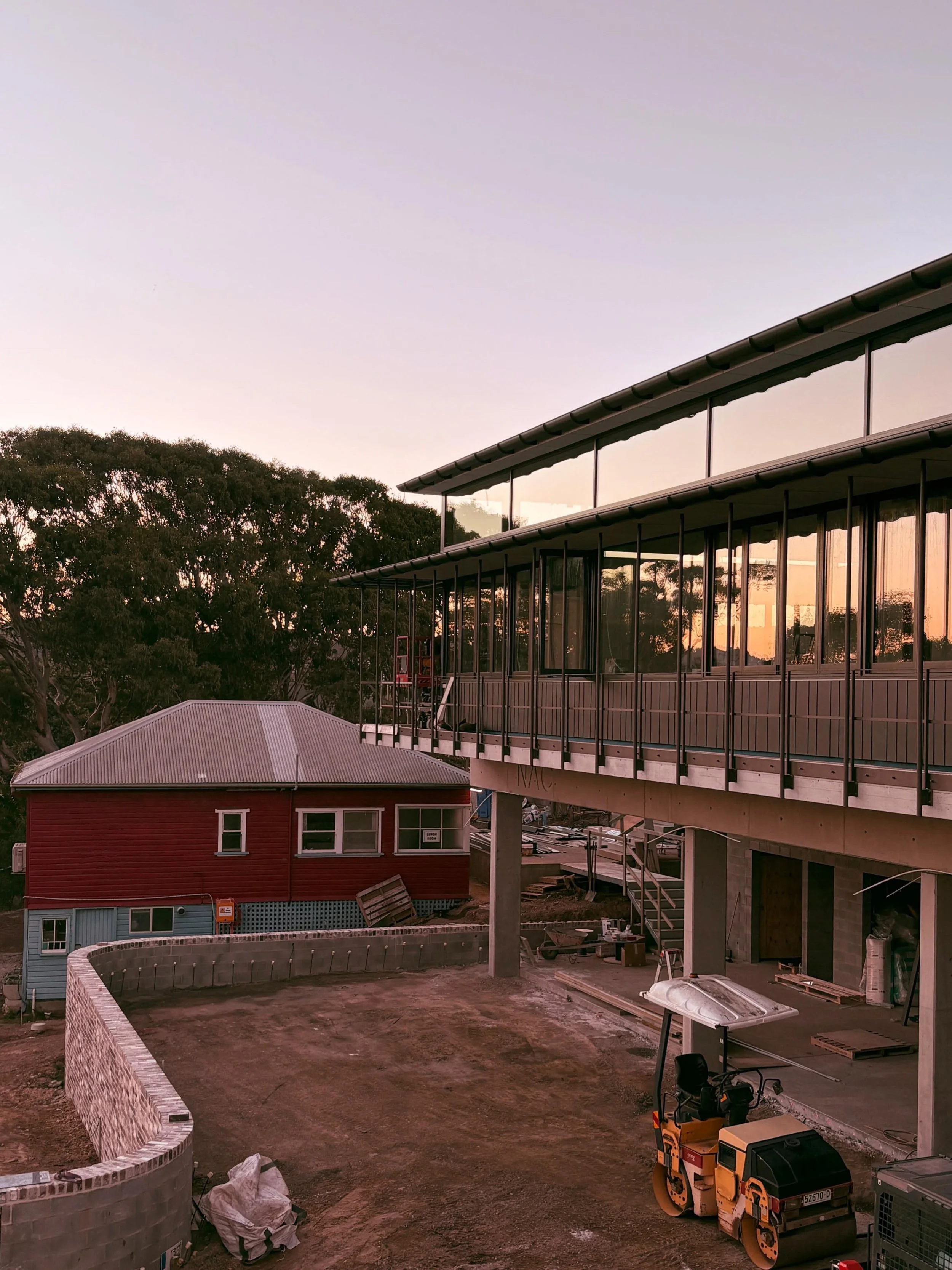 A construction site of Narooma School of Arts with a multi-story building in progress, surrounded by trees and an unfinished courtyard with construction equipment and materials. The school will open late 2026 and be the home of Mantra Flow classes