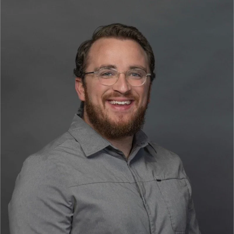 A young man with glasses, a beard, and brown hair smiling, wearing a gray button-up shirt, against a dark gray background.