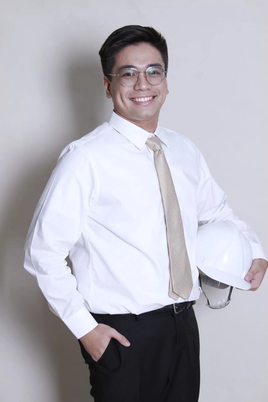 A smiling young man wearing glasses, a white dress shirt, and a light-colored tie, holding a white safety helmet in his left hand, standing against a plain off-white wall.