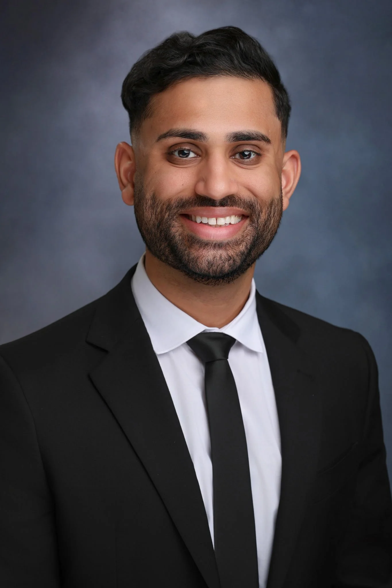A smiling man with dark hair and a beard, wearing a black tuxedo and black tie, against a dark gradient background.