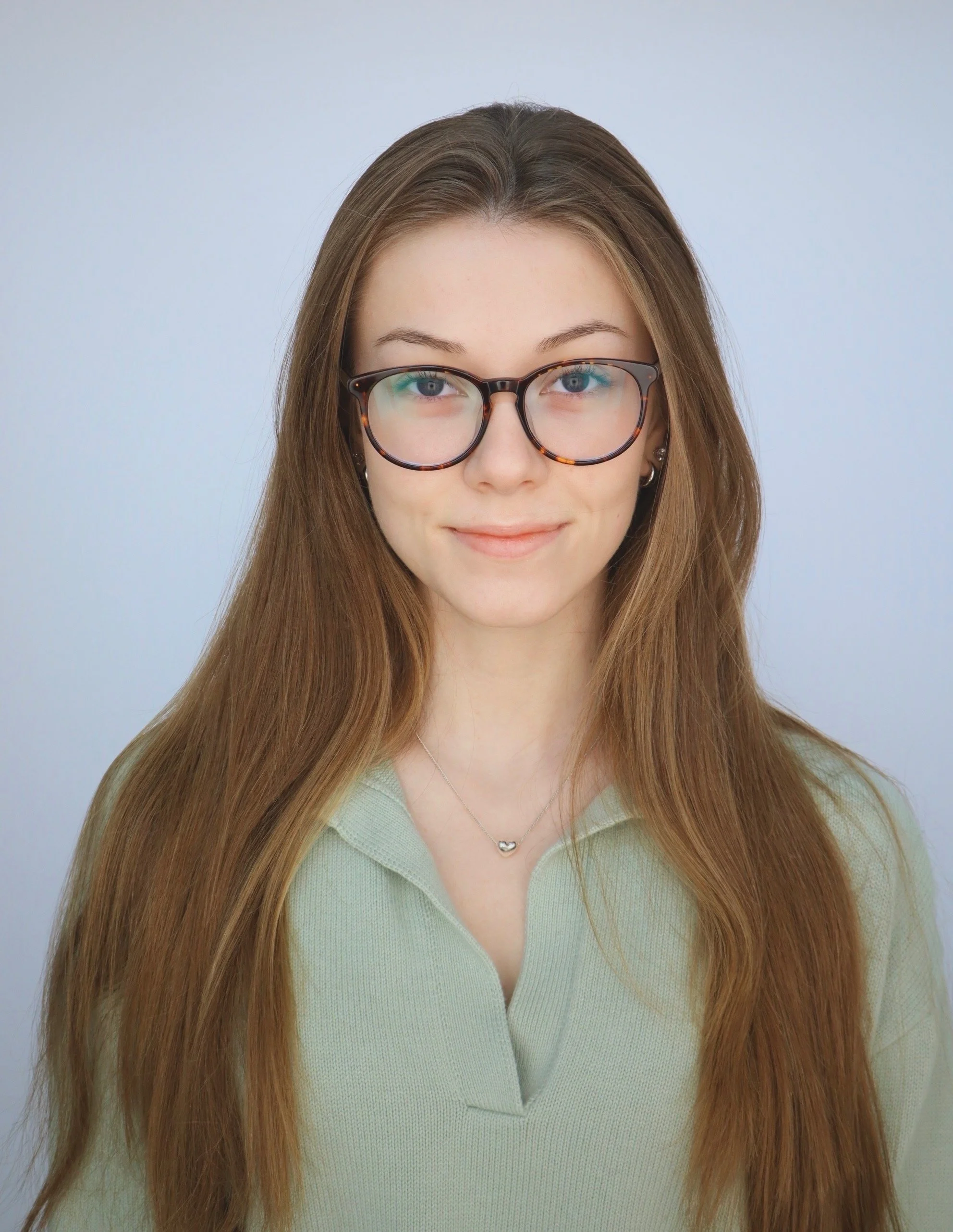 A young woman with long, straight, light brown hair and blue eyes wearing round tortoise shell glasses, a light green sweater, and a silver heart-shaped necklace, smiling softly at the camera against a plain light grey background.
