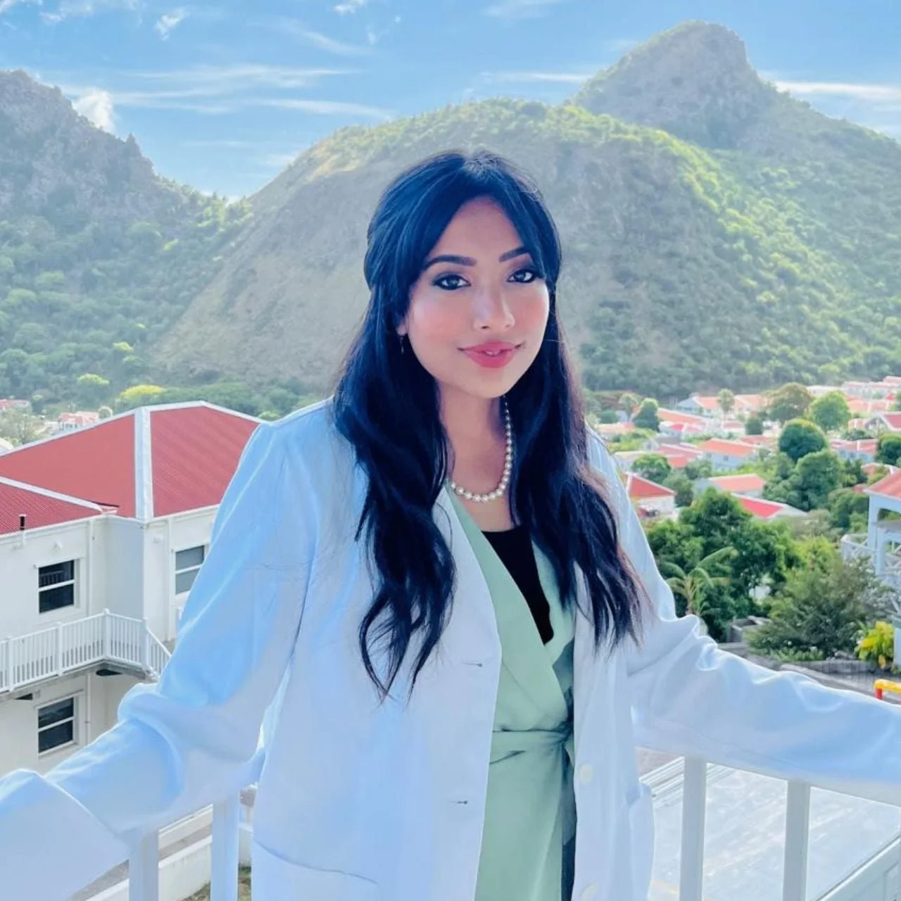 A woman with long black hair wearing a white blazer and pearl necklace, standing on a balcony with a scenic mountainous landscape in the background.