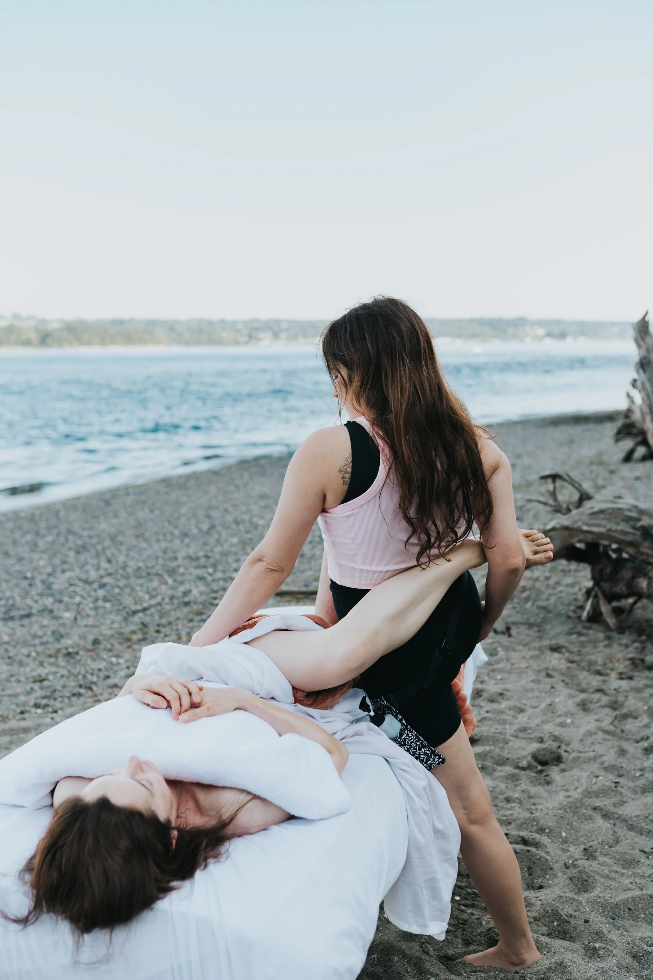 Two women practicing yoga on a beach, with one woman lying on a white towel and the other woman assisting in a pose, near the shoreline with water and distant land in the background.