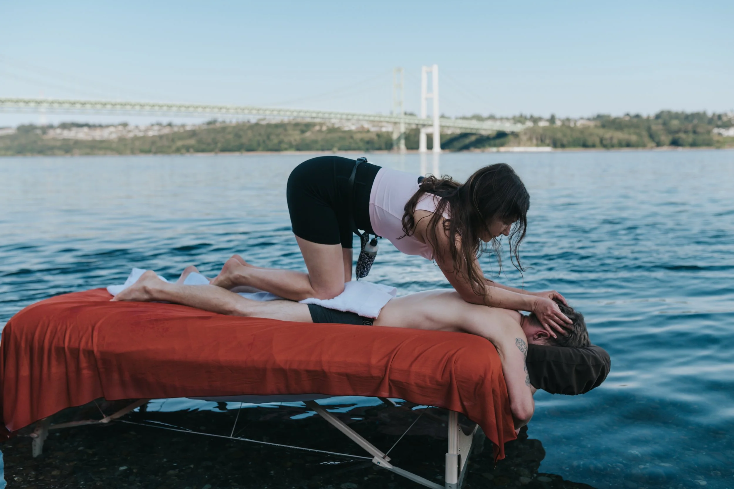 A person receiving a massage outdoors on a massage table by a body of water with a bridge in the background.