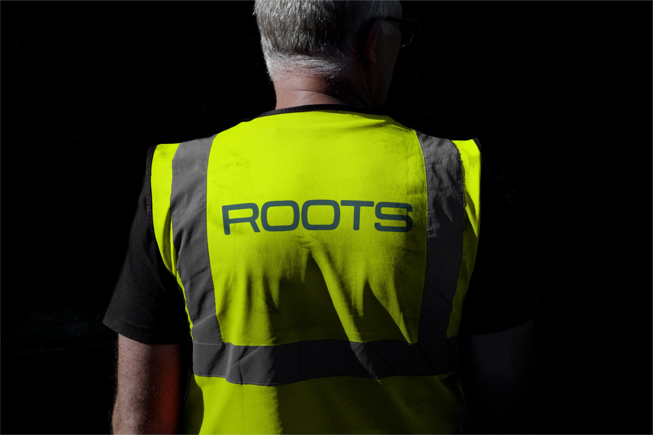 Person seen from behind wearing a yellow reflective vest with the word 'ROOTS' printed on the back, dark background.