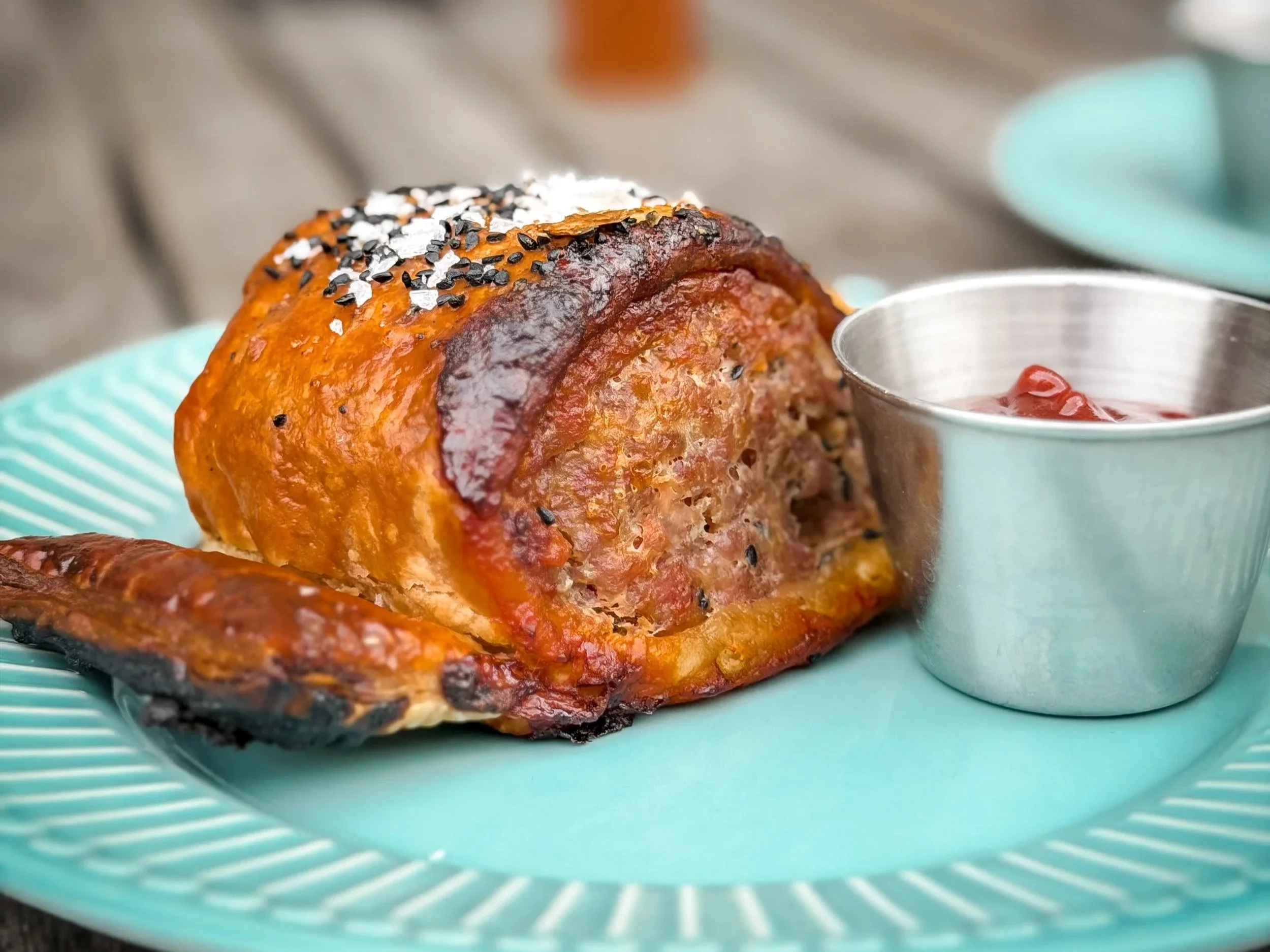 A baked salmon fillet topped with black sesame seeds and possibly white sesame seeds, served on a turquoise plate with a small cup of red dipping sauce on the side.