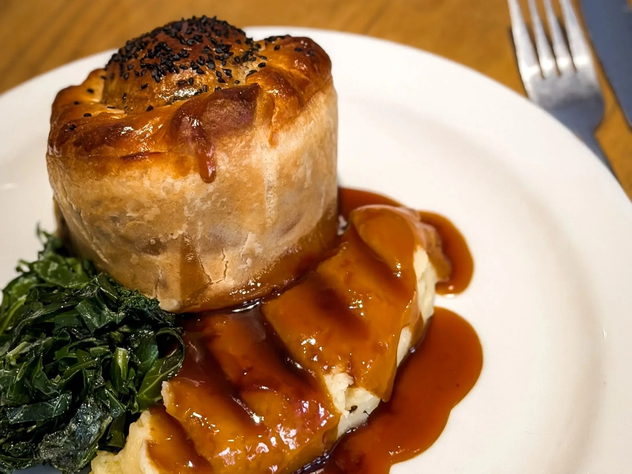 A plate with a serving of mashed potatoes topped with barbecue sauce, accompanied by cooked greens, and a baked stuffed chicken breast with melted cheese and black pepper on top.