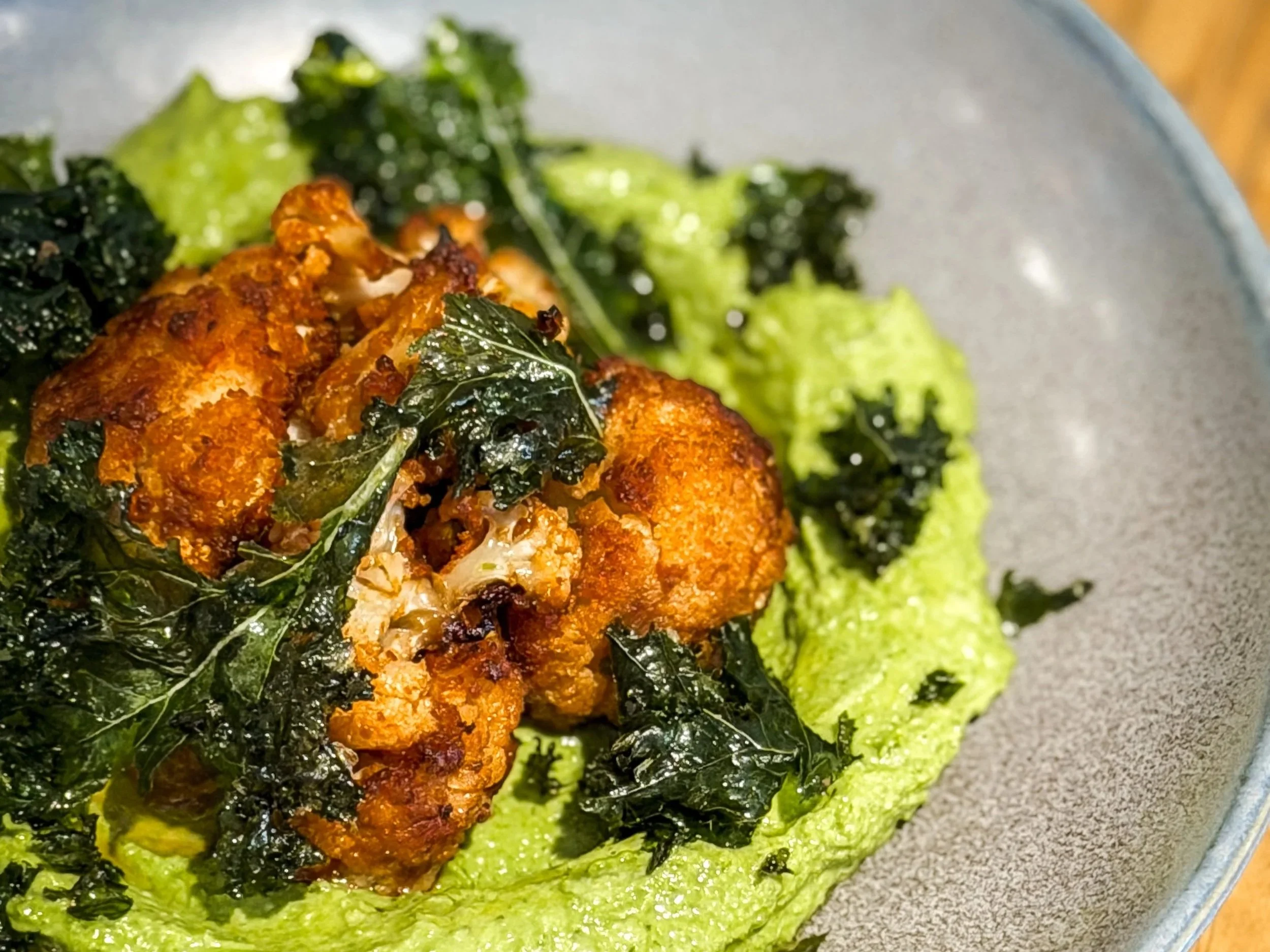 Close-up of roasted cauliflower coated in spices, topped with kale, on a gray plate with green mashed potatoes.